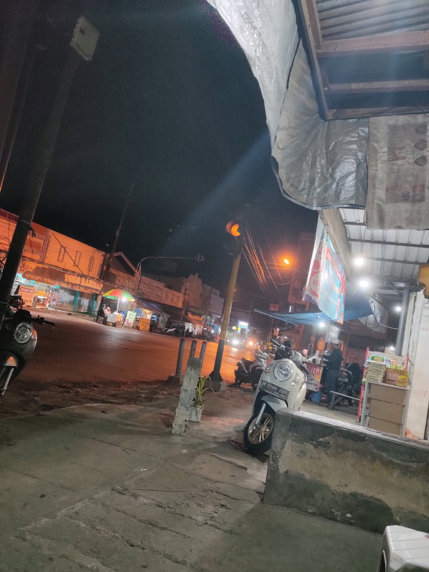 The image depicts a nighttime street scene in Codo, Indonesia. In the foreground, a light-colored scooter is parked on a concrete sidewalk. Beyond the scooter, the street is illuminated by streetlights and the lights from buildings. Several other motorcycles are parked along the street. In the midground, there are small food stalls with colorful umbrellas. One stall has a sign that appears to be in Indonesian. People are visible at the stalls. The buildings lining the street are lit up, and some have signage. A traffic light is visible on the left side of the image, showing a yellow light. The overall environment appears to be a commercial or residential area in a city at night.