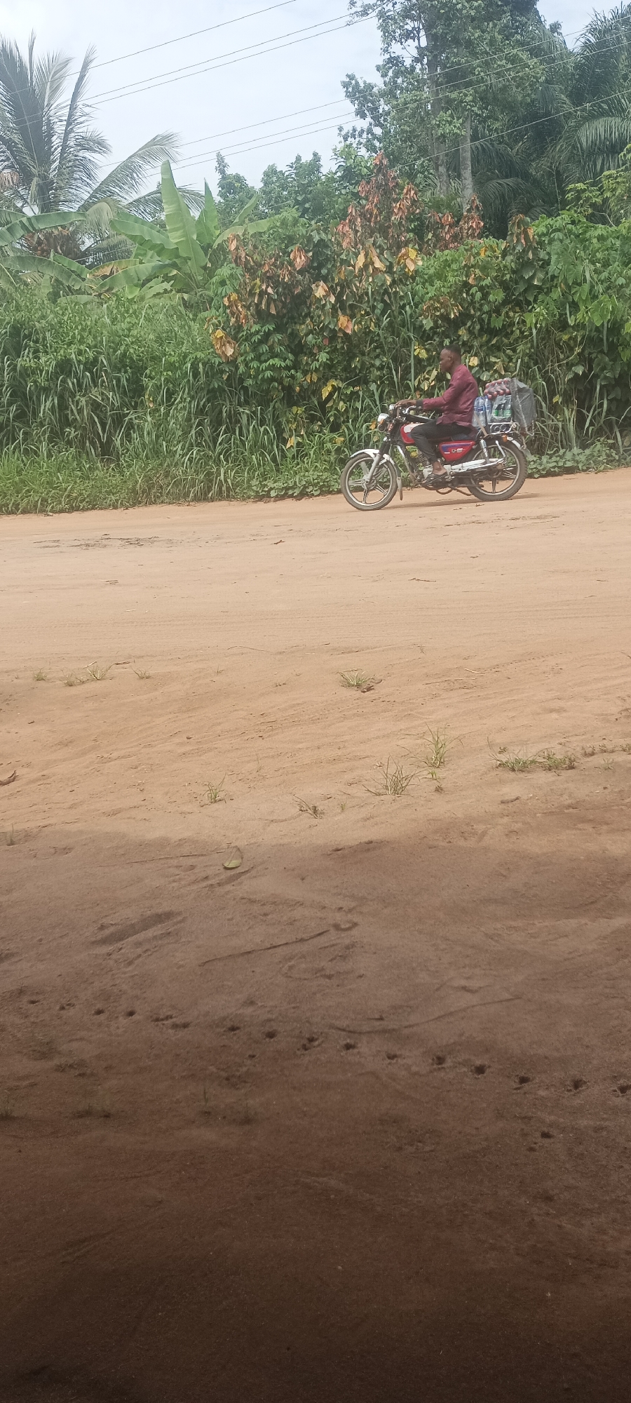A man rides a motorcycle loaded with goods on a dirt road in Amauzu, Nigeria. The road is unpaved and sandy. Lush greenery lines the side of the road. The sky is overcast. There are tracks in the dirt.