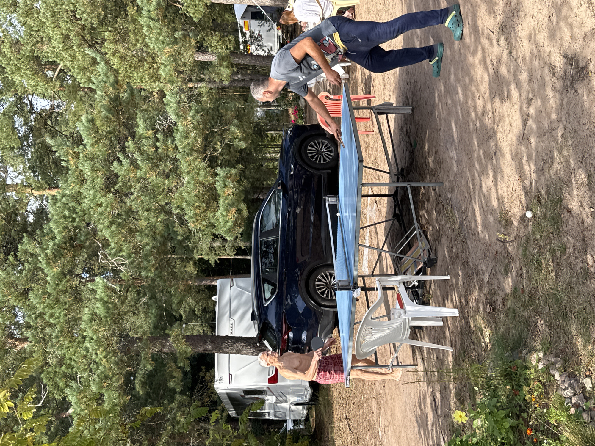 In Ozerki (Krasnodolinskaya), Russia, two men are playing table tennis outdoors on a sandy ground surrounded by trees. One man, wearing red shorts and shirtless, is positioned on the left side of the table. The other man, in a grey t-shirt and dark pants, stands on the right. A blue table tennis table is set up between them, with a white ball visible on the table. In the background, a dark blue SUV is parked next to a white caravan. Further behind, another white caravan and a lighter colored trailer are visible. The environment is characterized by dense pine trees and scattered sunlight on the ground.