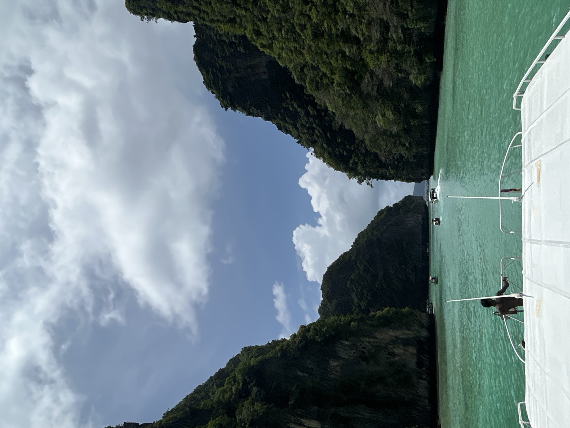 The image depicts a view from a boat's bow, looking forward into a narrow channel between steep, jungle-covered limestone karsts. The water is a vibrant turquoise and appears calm, with several long-tail boats and speedboats scattered in the distance. A person, presumably a crew member, stands near the railing on the left side of the boat's deck, facing away from the camera. The sky is partly cloudy, with a mix of white cumulus clouds and patches of blue. This scene is characteristic of the Maya Bay area near Ao Nang, Thailand, known for its dramatic karst formations and clear waters.