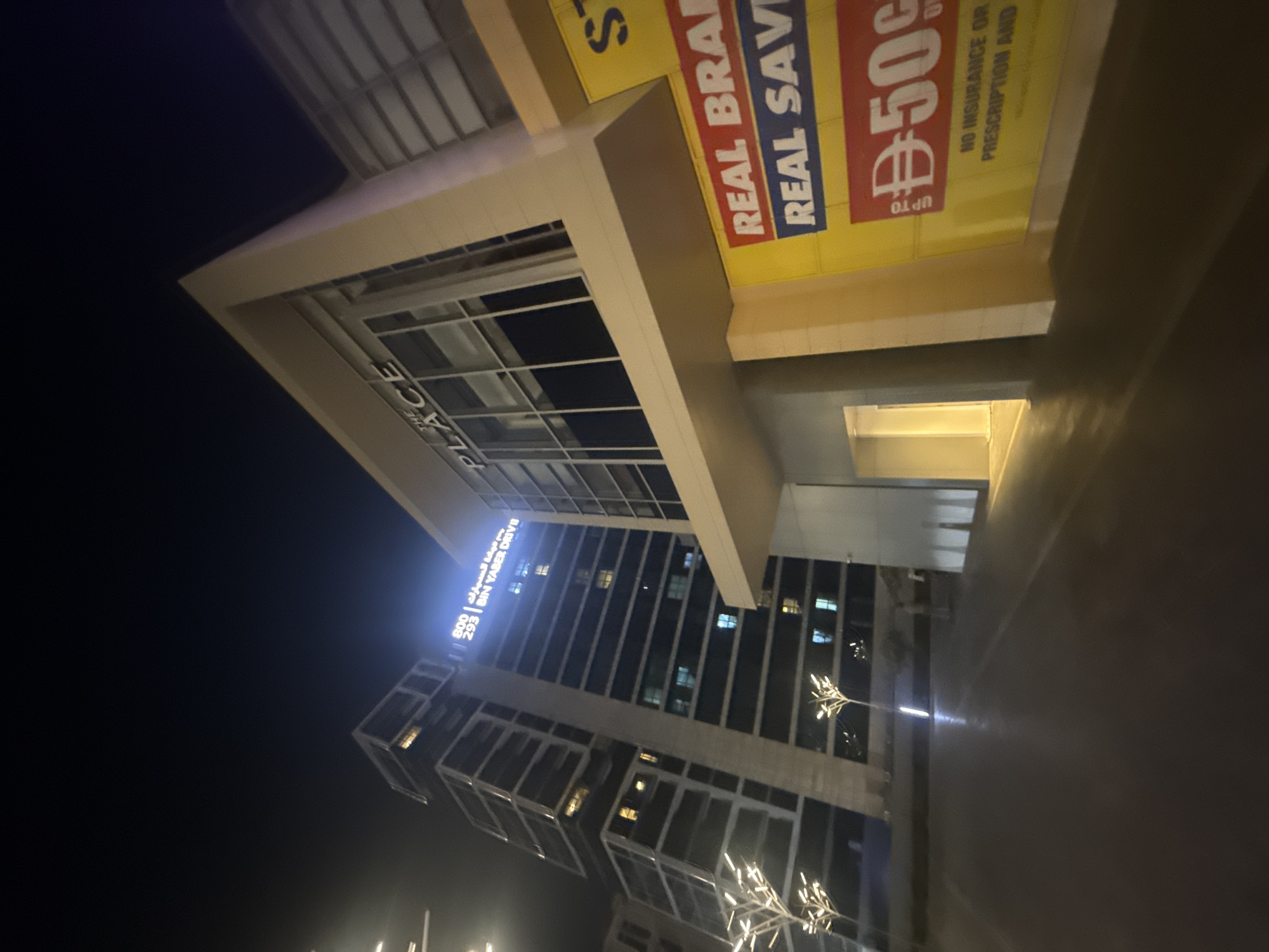 The image depicts a nighttime street-level view of a modern urban environment in Dubai, United Arab Emirates. In the foreground, a dark, wet-looking pavement reflects the ambient light. To the right, a large yellow advertisement is partially visible, featuring red lettering that reads "REAL BRA" and "REAL SAV," with a prominent red rectangle containing white text that appears to be "¥50c." Below this, more text in smaller print indicates "NO INSURANCE OR PRESCRIPTION AND."
Dominating the mid-ground and background are two distinct buildings. On the left, a tall, modern skyscraper with a dark facade and illuminated windows rises against the night sky. The skyscraper has a distinctive, angular design with many glass panels. The building on the right is also modern, with lighter-colored cladding and large windows. A prominent sign on this building displays the word "THE PLACE" in large white letters, with some Arabic script above it. The area between the buildings is illuminated by stylized, tree-like light fixtures with white branches. The overall scene suggests a commercial or mixed-use area, likely a retail or business district, characterized by contemporary architecture and nighttime illumination. There are no visible people in the immediate foreground or mid-ground, though a blurred figure may be present in the far background. The lighting is artificial, creating a dramatic effect with strong contrasts between illuminated areas and shadows.