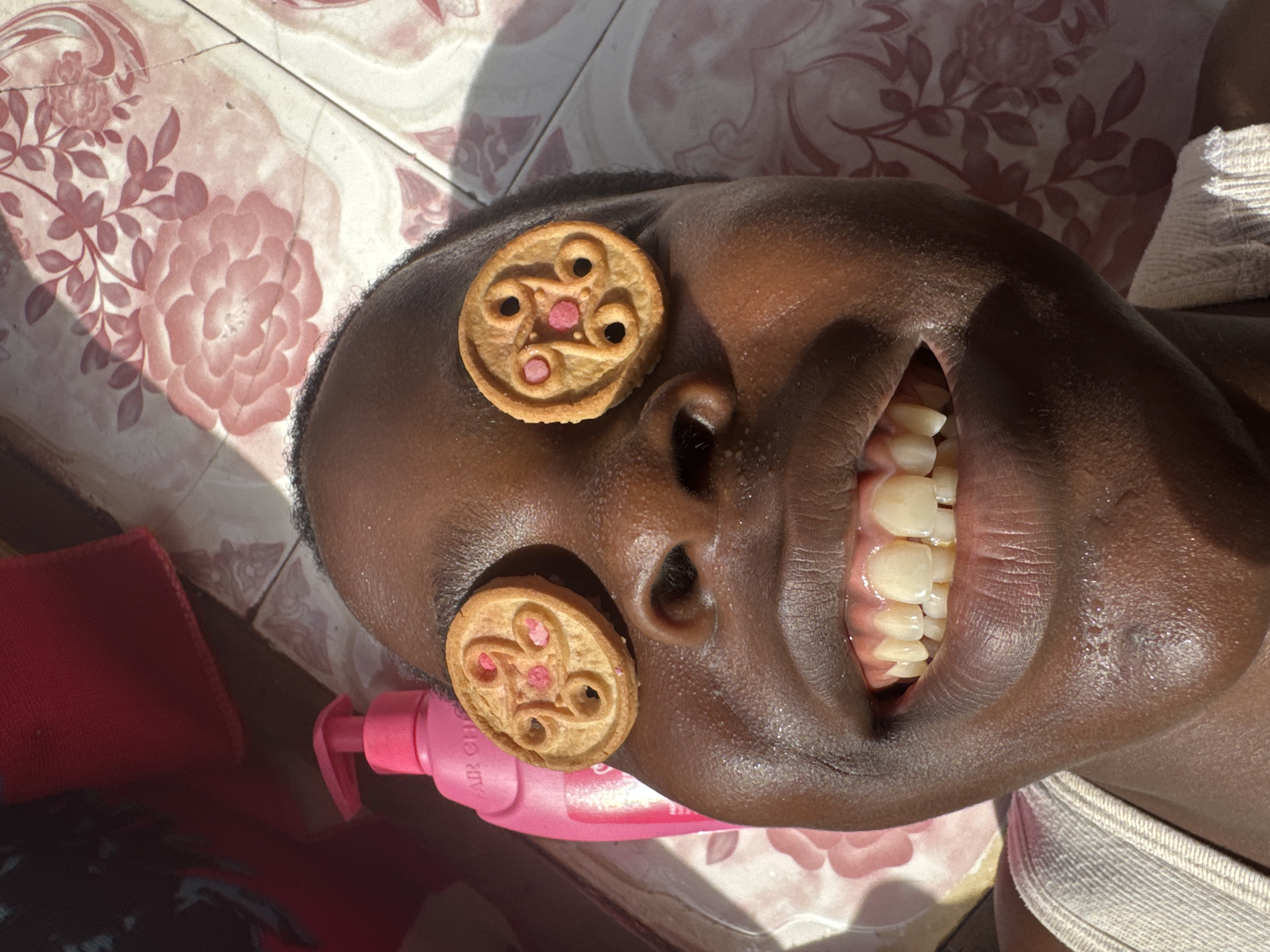 A medium shot shows a person, likely in Onitsha, Nigeria, lying on a surface covered with rose patterned floor tiles. A pink bottle of hair product is positioned near the person's left ear. Two round biscuits with a pink filling are placed over the person's eyes. The person is smiling, revealing their teeth. A partially visible red cloth is at the bottom left. The person is wearing a white garment. The scene is well-lit.