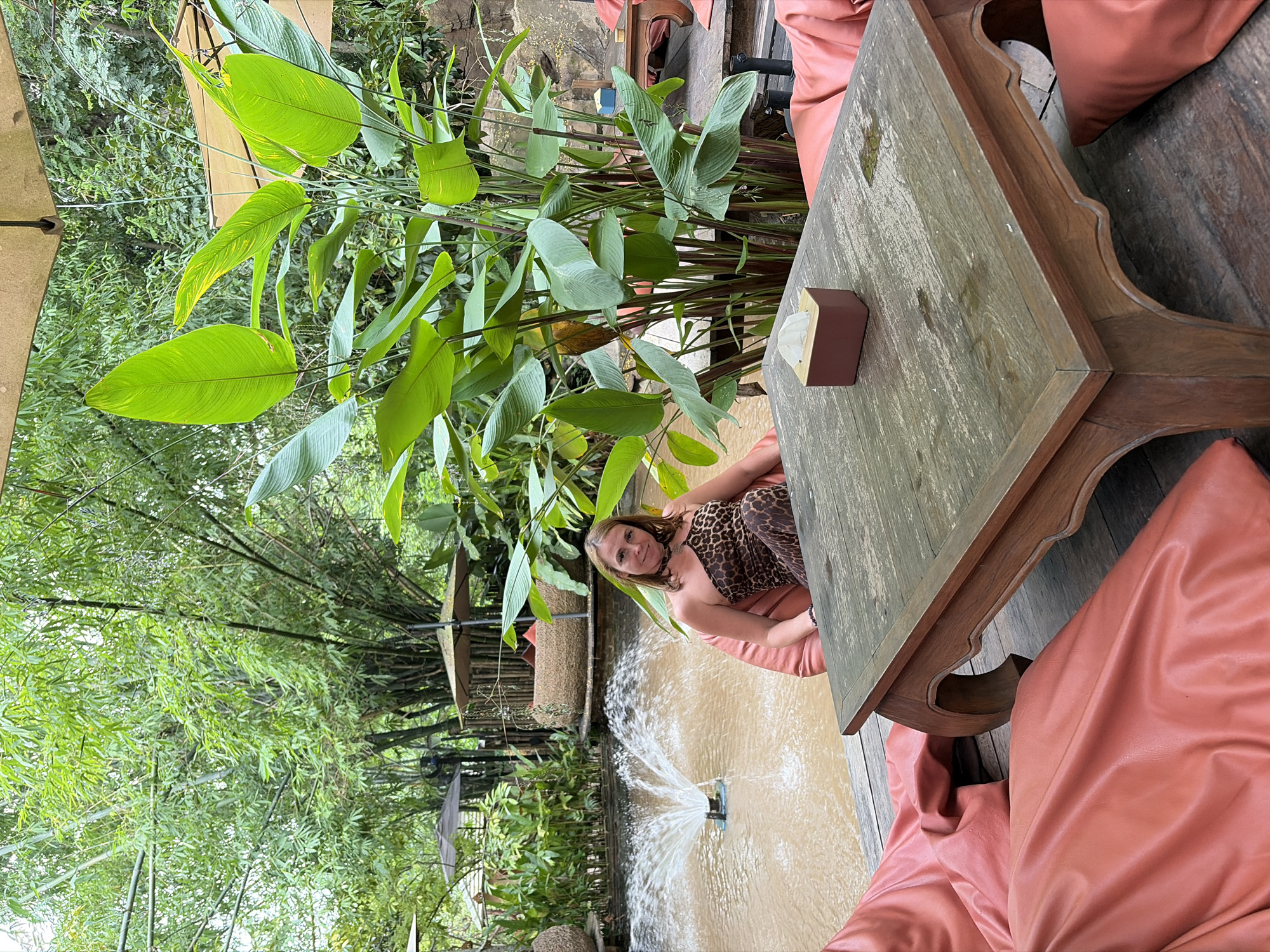 A woman is seated in a beanbag chair next to a wooden table and a fountain in Tambon Mae Raem, Thailand. She is wearing a leopard print one-shoulder jumpsuit and a choker necklace. The fountain is in a shallow pond surrounded by lush greenery and bamboo. The table is dark wood with ornate legs and has a tissue box on it. The beanbag chairs are a vibrant coral color and are scattered around the table. In the background, there are thatched umbrellas and seating areas, indicating an outdoor dining or lounge space. The overall setting appears to be a tropical resort or restaurant.