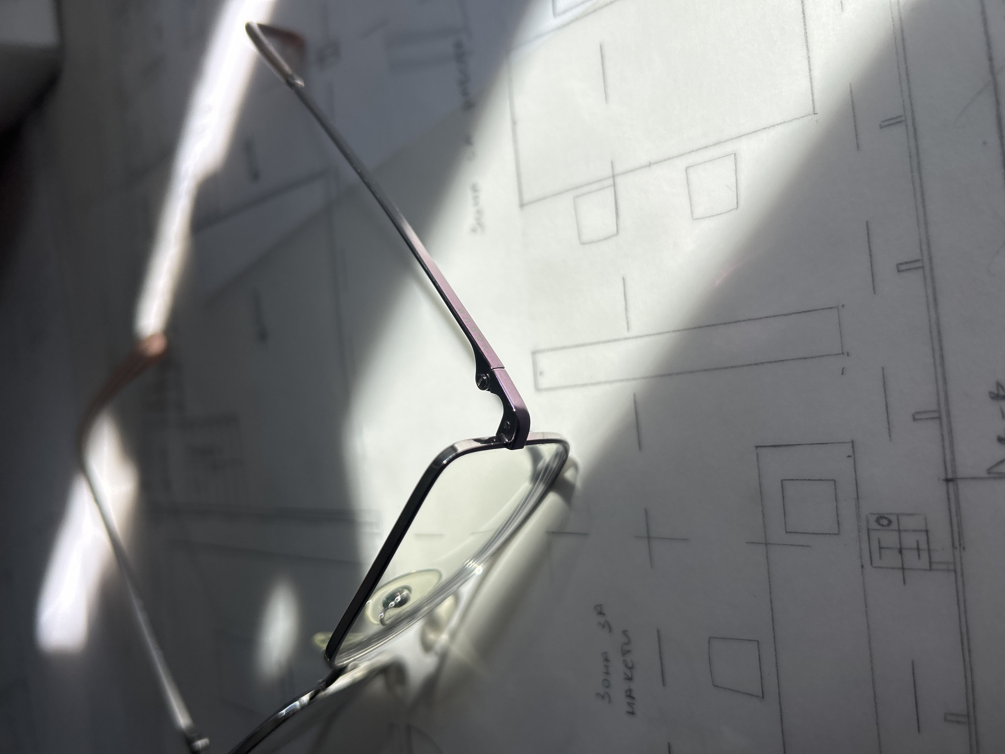 A pair of glasses rests on a piece of paper with technical drawings and handwritten notes. The glasses have a thin metal frame with a dark, possibly black or dark gray, color. The temple arm has a hint of purple. The paper below the glasses is white and covered with various geometric shapes and lines, suggesting a schematic or blueprint. Handwritten notes in a non-English language are also visible. The overall environment is brightly lit, with strong shadows cast by the glasses and other objects. The location is unknown, however text in Cyrillic is visible on the paper. The city of Plovdiv, Bulgaria is not visually confirmed and an assumption.