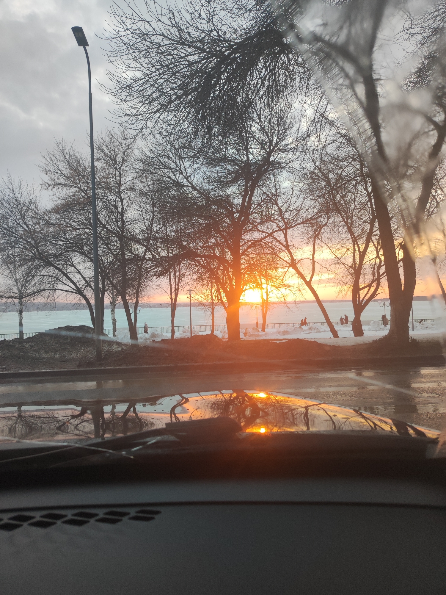 The image shows a view from inside a vehicle, looking out at a waterfront scene in Votkinsk, Russia. The scene is dominated by bare trees silhouetted against a sunset or sunrise. In the background is a body of water with ice. A fence runs along the water's edge, behind which several people are visible. A street lamp stands to the left of the trees. The foreground shows a wet surface reflecting the sunset. The vehicle's dashboard is visible at the bottom of the frame.
