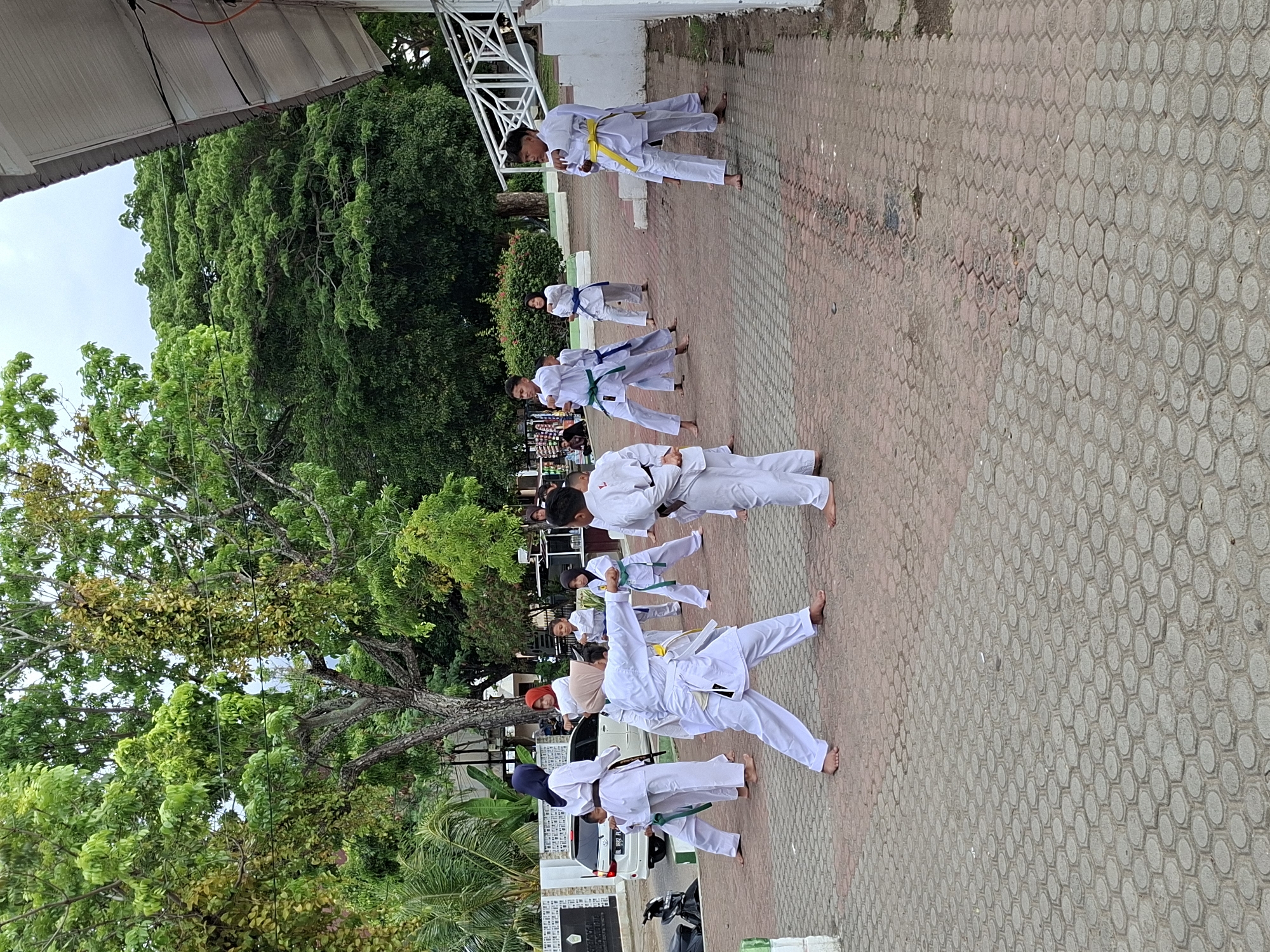 A group of people of varying ages are engaged in martial arts practice outdoors. They are wearing white martial arts uniforms with different colored belts, suggesting varying skill levels. The practice is taking place on a paved area constructed of interlocking hexagonal pavers, which is next to a building and under the shade of large trees. The location appears to be in Lhokseumawe, Indonesia, indicated by the car license plate starting with "BL". Other details in the frame include parked vehicles (a scooter and a car), parts of buildings, trees, and vegetation.