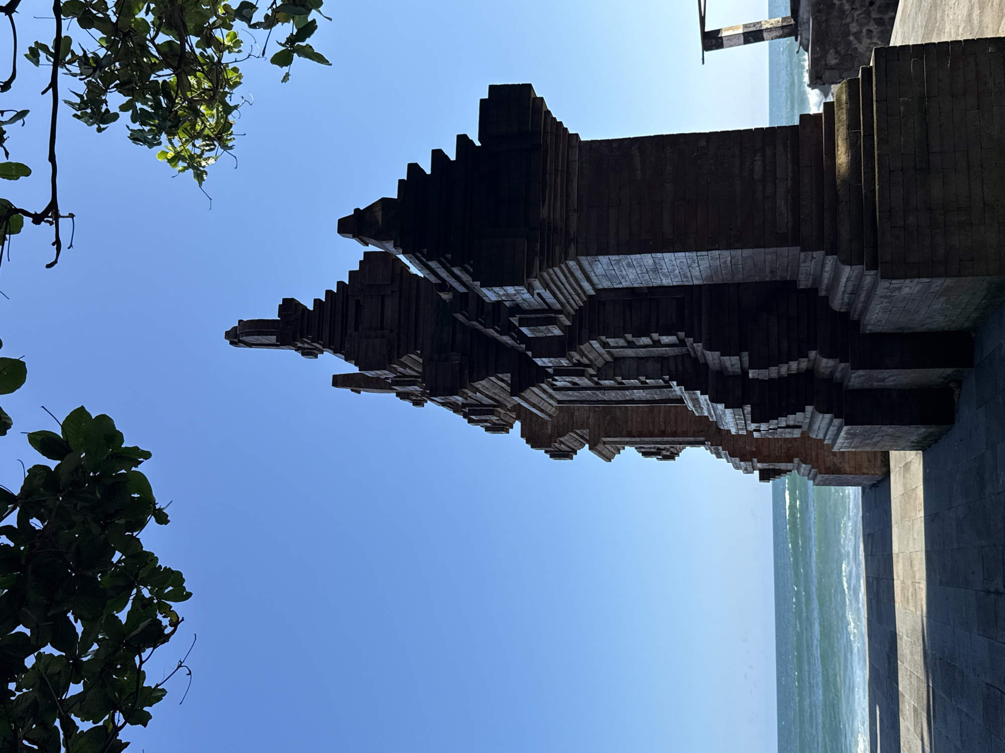 The image, taken from a low angle, depicts a structure, most likely a temple, silhouetted against a clear, bright blue sky. Some green foliage is visible on the upper left, suggesting the presence of trees. The structure, built with brick-like elements, has a multi-tiered, stepped design. A portion of the ocean and a stone wall are visible on the right side. This is likely a coastal temple in Mengwi, Indonesia.
