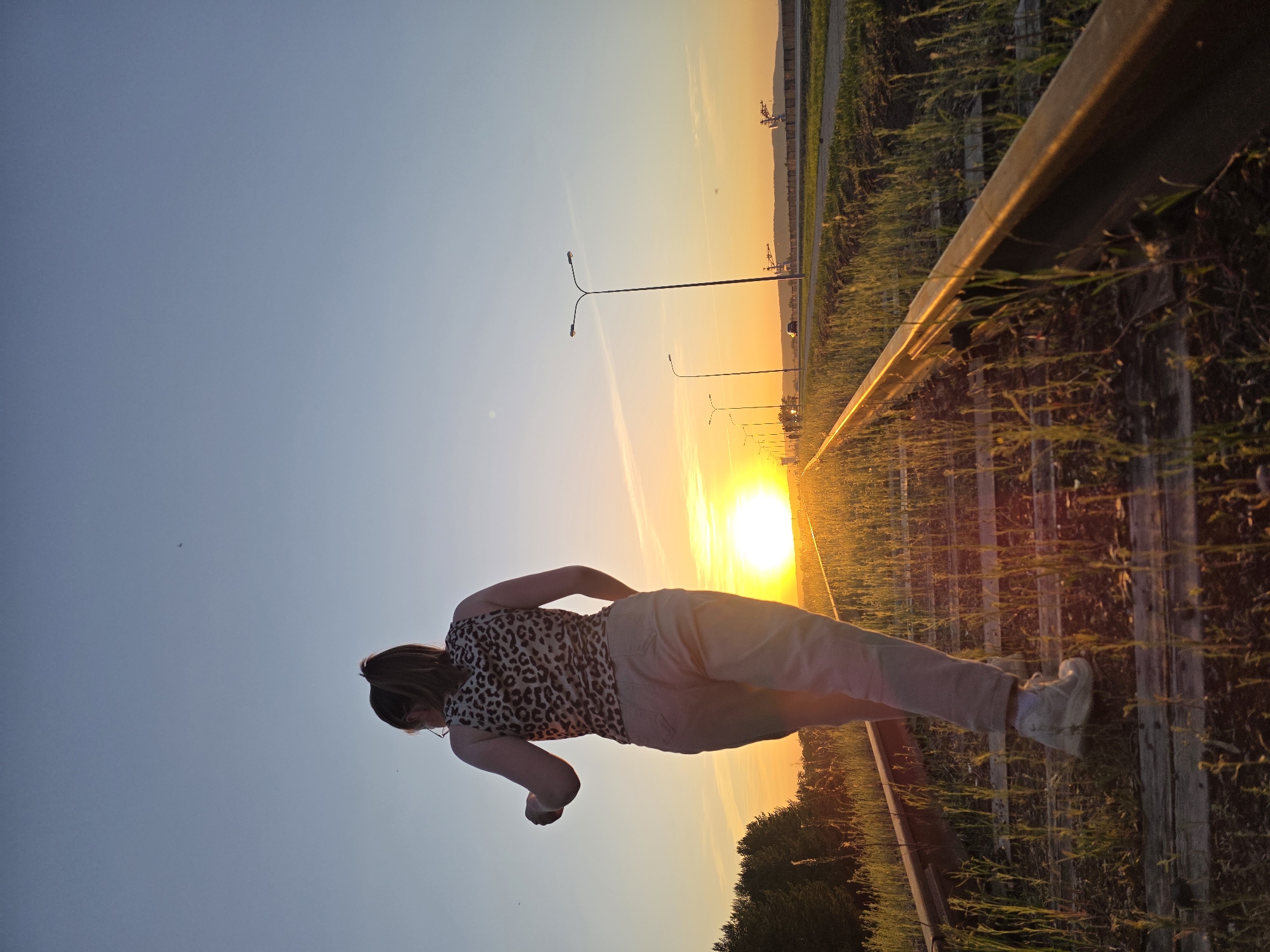 A woman stands on a set of railway tracks in the city of Chaikovsky, Russia, during a sunset. She wears a leopard-print top, light-colored pants, and white sneakers. Her hair is pulled back. She is seen from behind. The railway tracks stretch into the distance towards the setting sun, which is visible between the tracks. Street lights line a road that runs parallel to the railway tracks on the right. There are trees and bushes along the edges of the tracks and road. The sky is blue on the left and transitions to orange and yellow near the horizon.