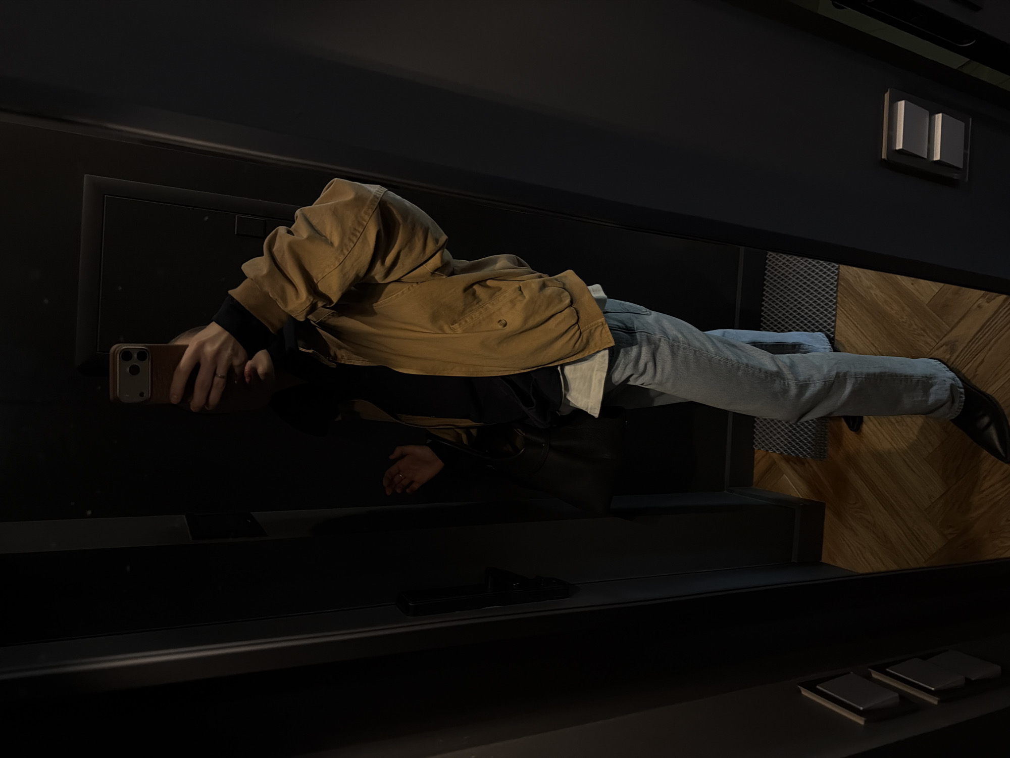 The image, rotated 90 degrees clockwise, depicts a person taking a mirror selfie in a modern indoor setting, likely in Moscow, Russia. The individual is positioned centrally, holding a smartphone with a light brown case and a triple camera setup, which obscures their face. They are wearing a light brown or tan jacket, light blue jeans, and a dark long-sleeved shirt underneath, with a white layer visible at the waist. A ring is worn on one of their fingers, and a black shoe is visible on the right foot.
The surroundings feature dark, possibly black or very dark grey, walls and surfaces, contributing to a sleek, contemporary aesthetic. On the right side of the frame, there is a section of warm-toned wooden paneling arranged in a chevron or herringbone pattern. Adjacent to this, a rectangular panel with a dark, textured, possibly gridded surface is visible. Mounted on the dark wall are two modern, rectangular light switches. Below the person, a dark ledge or display surface holds two small, square, grey objects. The lighting is somewhat dim, casting subtle shadows and enhancing the modern atmosphere. No visible text is present in the image.