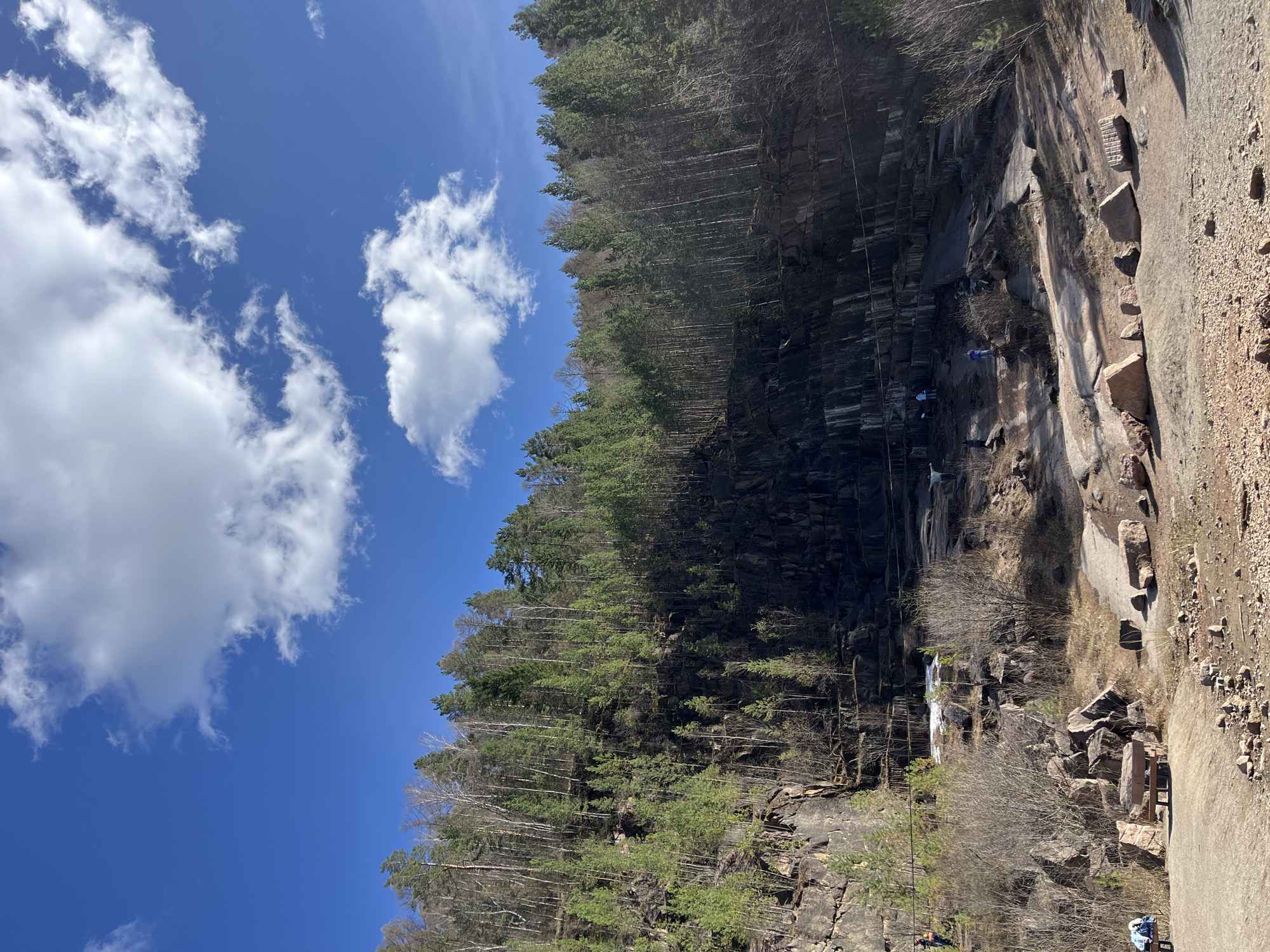A landscape photo shows a high rocky cliff and a blue sky with white clouds, likely in Krasnoyarsk, Russia. The cliff, located on the right side of the image, has varied rock types and surfaces, a few bushes, and sparse vegetation growing at the bottom. On the top of the cliff, there's a thick forest of evergreen and deciduous trees. A small waterfall is visible cascading down the cliff face. There are a few people climbing a zipline down the cliff. On the left side of the image, a clear blue sky is partially covered with puffy white clouds. The scene is likely a natural rock formation near or in the city of Krasnoyarsk.