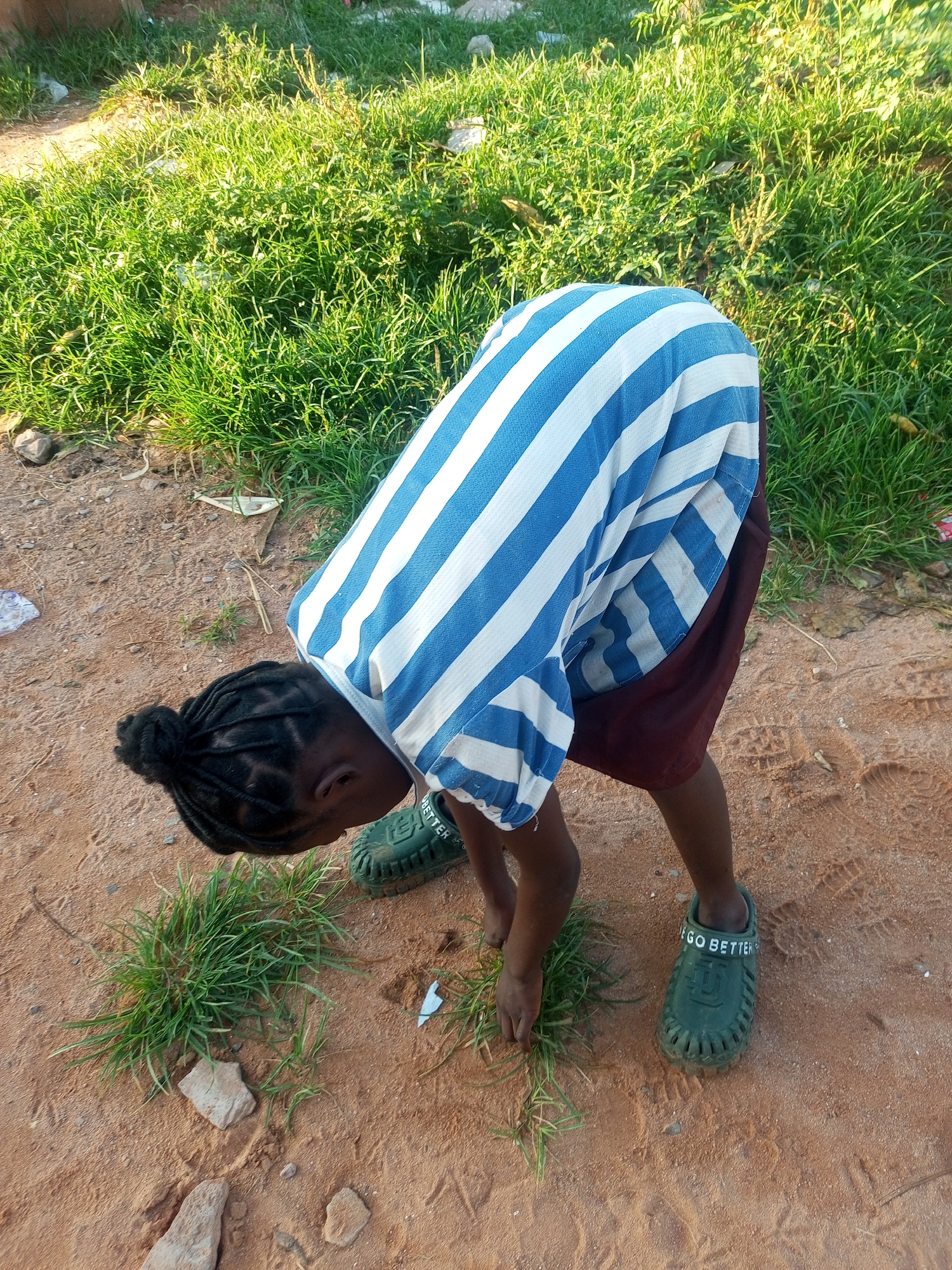 A young person, dressed in a blue and white striped shirt and a maroon skirt, is bent over in a dirt area with patches of grass and small stones. The person's head is bowed, and their hands are near a clump of grass on the ground. They are wearing dark green clogs with the words "GO BETTER" visible on the side. The background shows more grass and dirt. The scene appears to be outdoors in a rural or semi-rural setting within the city of Tempure, Nigeria.