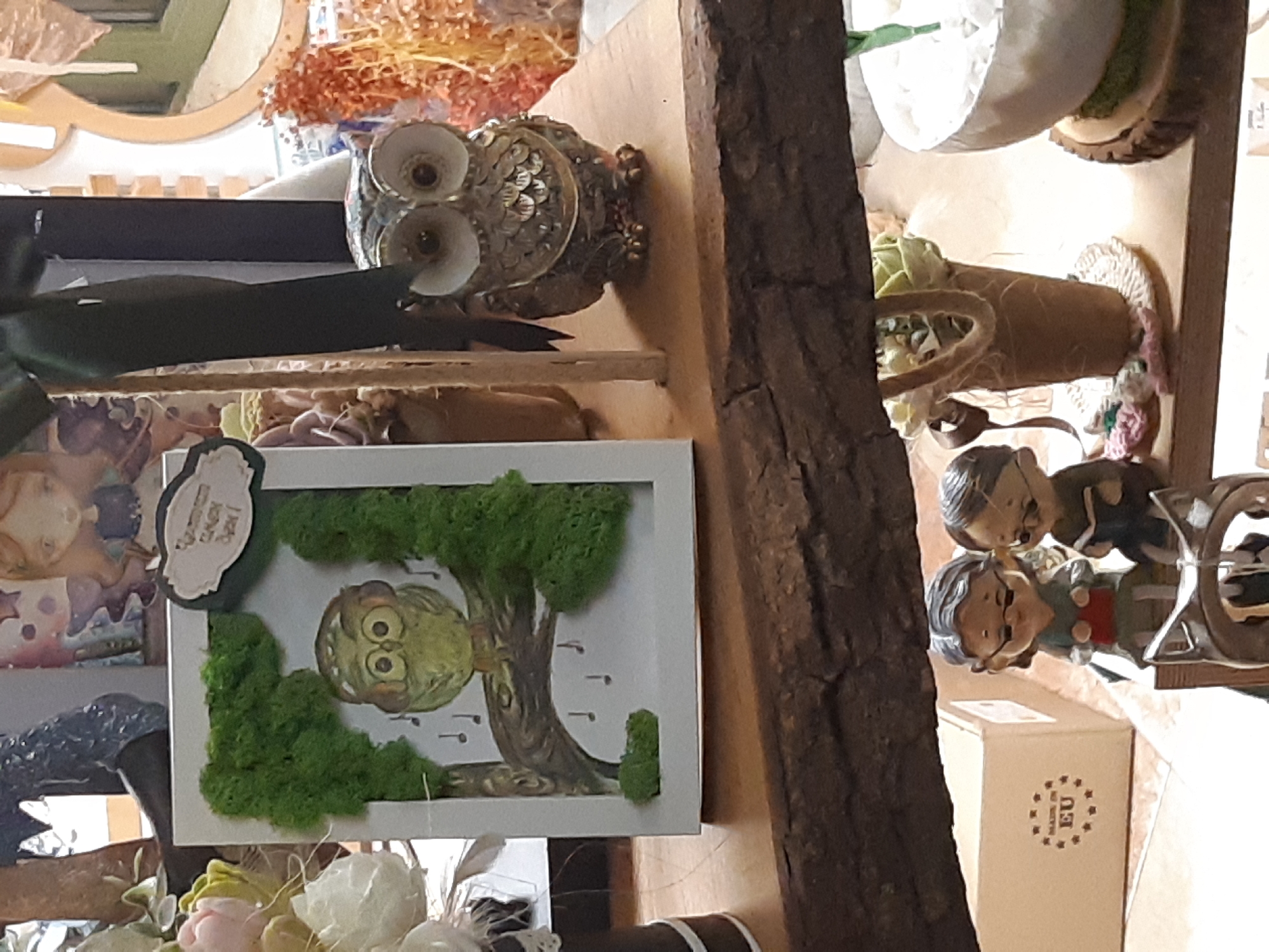 The image depicts a collection of decorative items displayed on a shelf, possibly in a shop in Plovdiv, Bulgaria.
Objects present include:
* A framed picture with a stylized owl made of moss.
* A decorative metal owl figurine.
* Small figurines of elderly people.
* A small box with a "Made in EU" logo.
* Other decorative items, including flowers and pots.
* A piece of tree bark serves as a vertical element within the display.
The general impression is that of a curated collection of crafts for sale. The background details are obscured but suggest a retail setting.