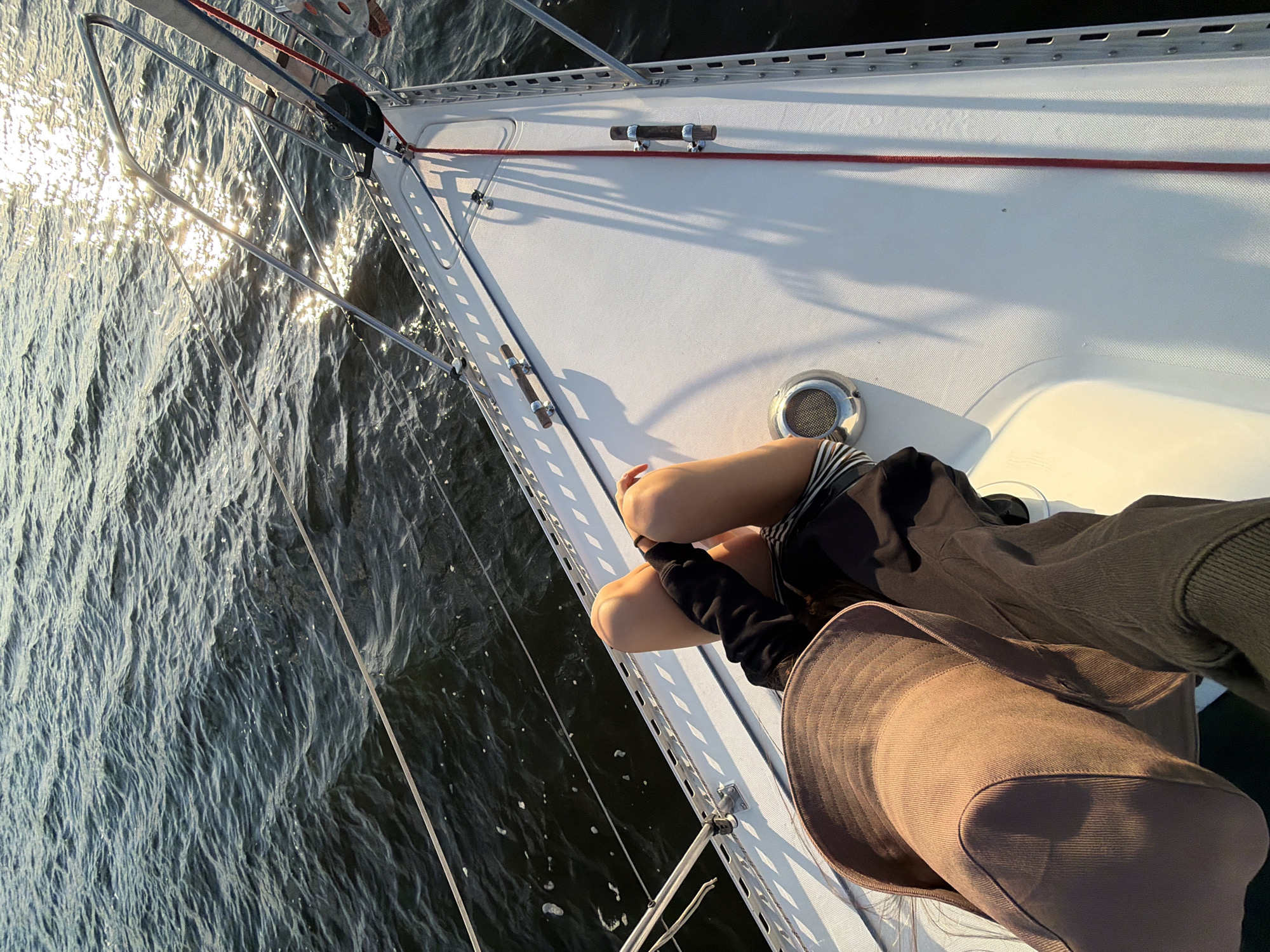 A first-person perspective shot shows a person sitting on the bow of a sailboat, with their legs extended forward. They are wearing a brown hat and a black sweatshirt. The water is dark blue and slightly choppy, with sunlight reflecting off the surface. The bow of the boat is white and clean, with several metal cleats and ropes visible. The scene appears to be a leisure activity on a body of water, likely a river or a bay. The specific location in Kamyshin, Russia, is not identifiable from the visual elements of the image.