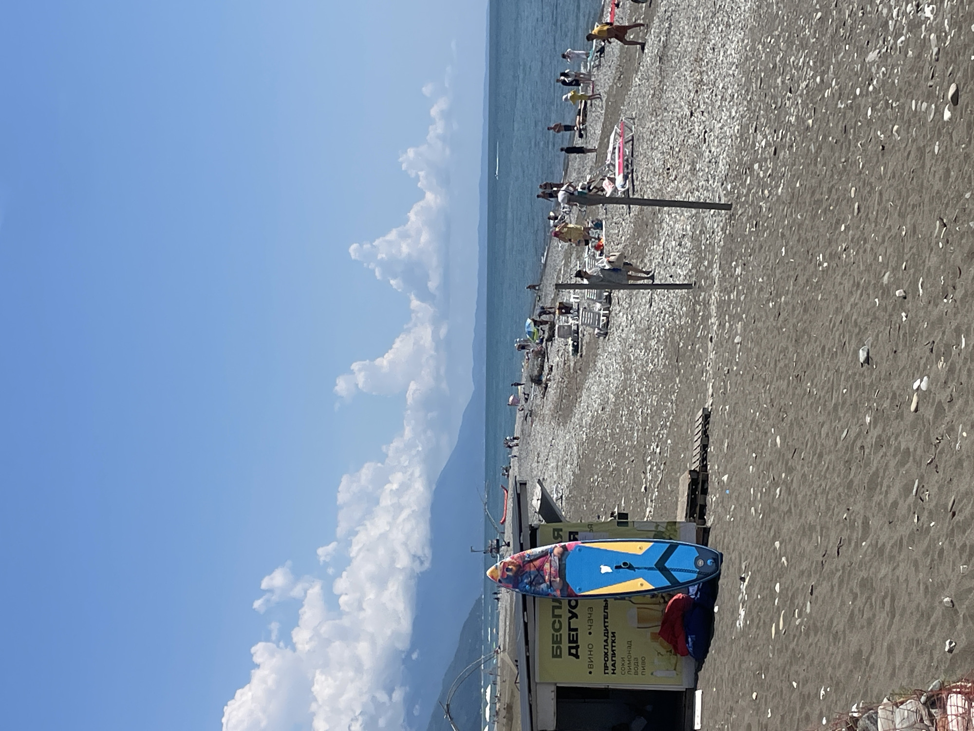 The image captures a sunny day at a pebble beach in Gagra, Abkhazia. The scene is dominated by the vast blue sea meeting a wide stretch of dark grey pebbles interspersed with dark sand. In the foreground, a colorful stand-up paddleboard is propped upright against a small kiosk. The kiosk has a yellow sign with Cyrillic text, advertising "FREE TASTING" and listing various beverages like wine, tea, juices, lemonade, water, and beer. Behind the kiosk, red and blue beanbag chairs are visible.
Further down the beach, numerous people are scattered, engaging in various beach activities. Some are lying on lounge chairs, others are standing or walking, and a few are in the water. The water is a deep blue-green, with small waves lapping the shore. In the distance, a hazy mountain range forms the backdrop, with a clear blue sky dotted with fluffy white clouds overhead. The overall impression is a typical summer day at a seaside resort, with a mix of leisure activities and commercial offerings.