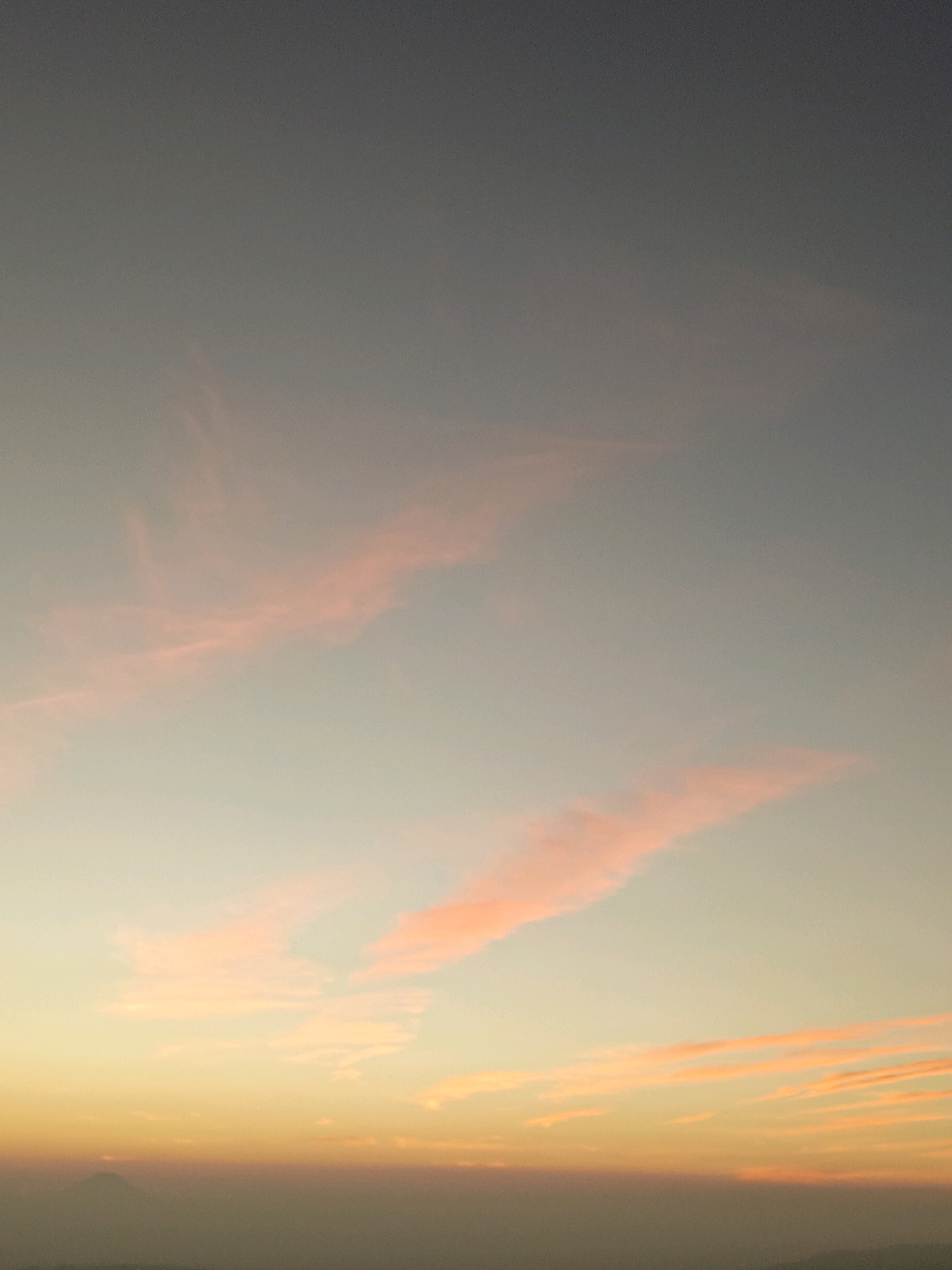 A photograph captures a serene dusk sky over Butuh Kidul, Indonesia. The sky transitions from a soft grey at the top to a warm gradient of peach and orange near the horizon. Wispy clouds, tinged with pink, are scattered across the upper and middle sections of the sky. In the distance, a faint silhouette of a mountain peak is visible on the left, emerging from a hazy, muted landscape that marks the transition to the earth. The overall atmosphere is tranquil and atmospheric, characteristic of a sunset or sunrise.
