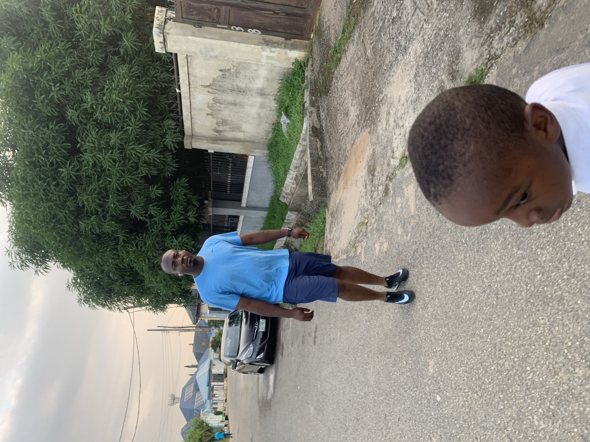 In Abuja, Nigeria, an outdoor scene captures a man walking on a paved road in a residential area. He is wearing a blue t-shirt and navy blue shorts, paired with black athletic shoes. Behind him, a dark-colored sedan is parked on the left side of the road. The environment features a lush green tree on the right, bordering a beige-colored wall with a metal gate. The sky is overcast, suggesting a cloudy day. In the foreground, a young child's head and shoulders are visible, facing to the left, partially out of focus. The child is wearing a white shirt. The street is lined with houses and a concrete drainage system.