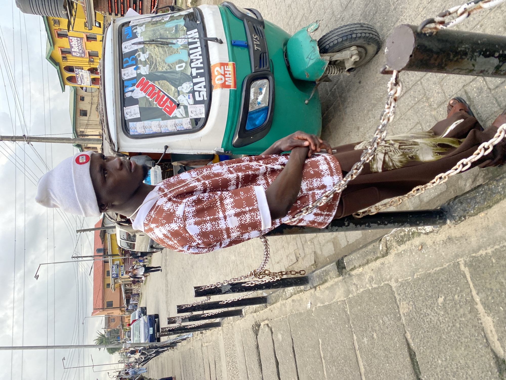 A young Black man wearing a white beanie and a brown and white patterned shirt stands leaning against a metal chain fence on a city street. The man is facing to his right, looking away from the camera, with his hands clasped in front of him. He is wearing brown pants and sandals. To his right is a green and white auto-rickshaw with various stickers on its windshield, including a red "RAHIL" sticker. In the background, a street scene unfolds with other vehicles, buildings, and pedestrians. The street appears to be paved with concrete and lined with a curb. The sky is overcast.