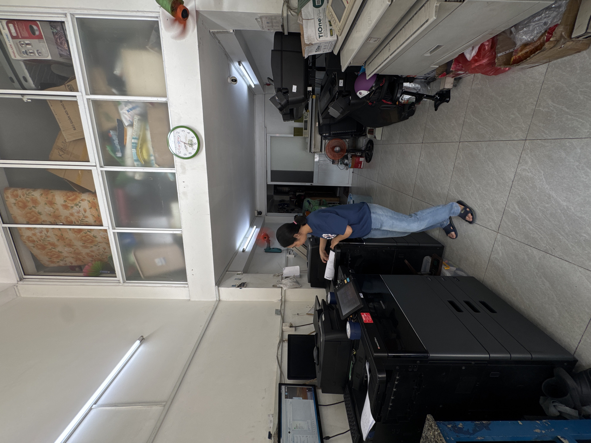 The image shows an interior view of what appears to be a copy and print shop. A woman in a blue t-shirt and jeans is standing in front of a large black Toshiba copier and printer. She is holding a document and looking at the copier's touchscreen. To her left are two computer monitors, a keyboard, and a printer. In the background, there are more printers, shelving units with various items, and a circular clock on the wall. The room is brightly lit by fluorescent lights. The overall environment suggests a functional workspace with an emphasis on office equipment.