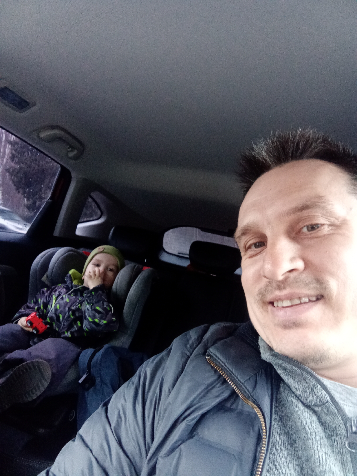 The image shows a man and a child in the back seat of a car. The man is in the foreground and appears to be taking a selfie. He is wearing a grey quilted jacket over a grey top. The child is in a grey car seat behind him, wearing a patterned jacket and a green hat, and holding a red toy. The background shows a window with a view of a snowy or wet street. Based on the location information (Izhevsk, Russia), the environment is likely cold and potentially snowy or wet. The overall impression is a casual moment captured inside a car during potentially inclement weather.