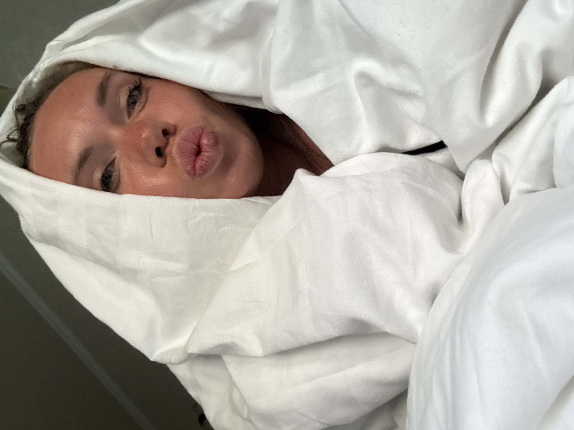 A woman with blond hair, wearing a white blanket, poses for a selfie in what appears to be a bedroom in Anapa, Russia. She is puckering her lips and looking at the camera. The blanket is draped over her head and shoulders, obscuring most of her body. The lighting suggests it is daytime.