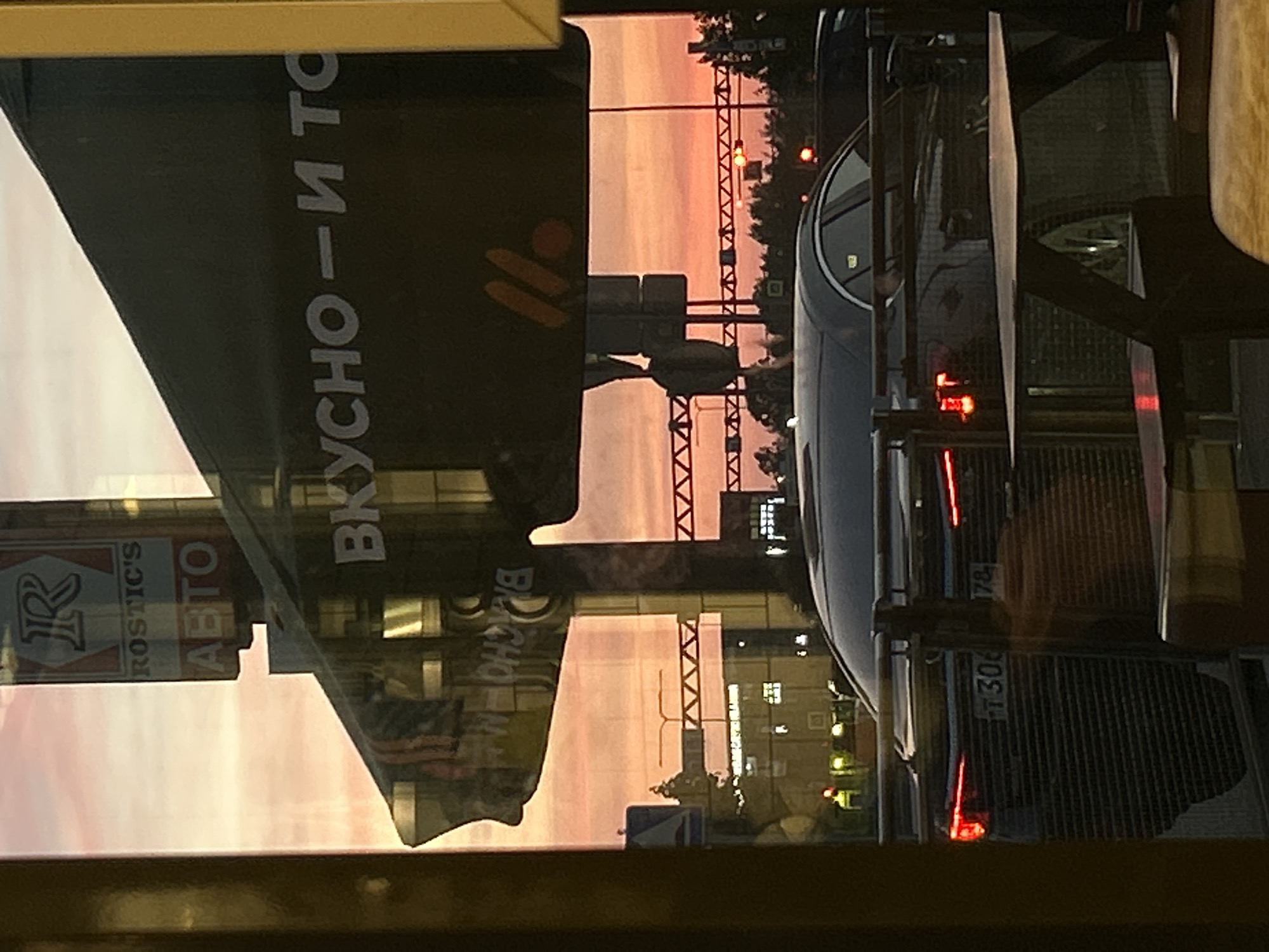 The image shows a view from inside a building, looking out onto a street in Saint Petersburg, Russia. A "Vkusno i tochka" sign (a Russian fast-food chain that replaced McDonald's) is visible in the reflection of the window. A "Rostic's" sign (a KFC franchise) is also reflected. Outside, there's a car with a license plate that reads "T 306 178", and traffic lights are visible. The sky appears to be at dusk or dawn, with a pinkish hue. The street scene includes buildings and trees.
