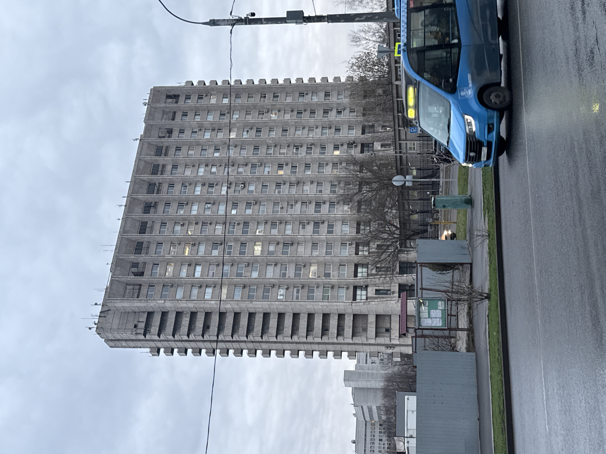 The image depicts a multi-story gray apartment building in Moscow, Russia. The building has a distinctive, brutalist architectural style, characterized by its rectangular shape, repetitive window pattern, and protruding concrete ledges. A blue bus is visible in the foreground, partially obstructing the view of the building. The environment is overcast and the road appears wet, suggesting recent rain. There are some bus stop structures, signs, trees and other buildings in the vicinity. There are no visible people in the shot.