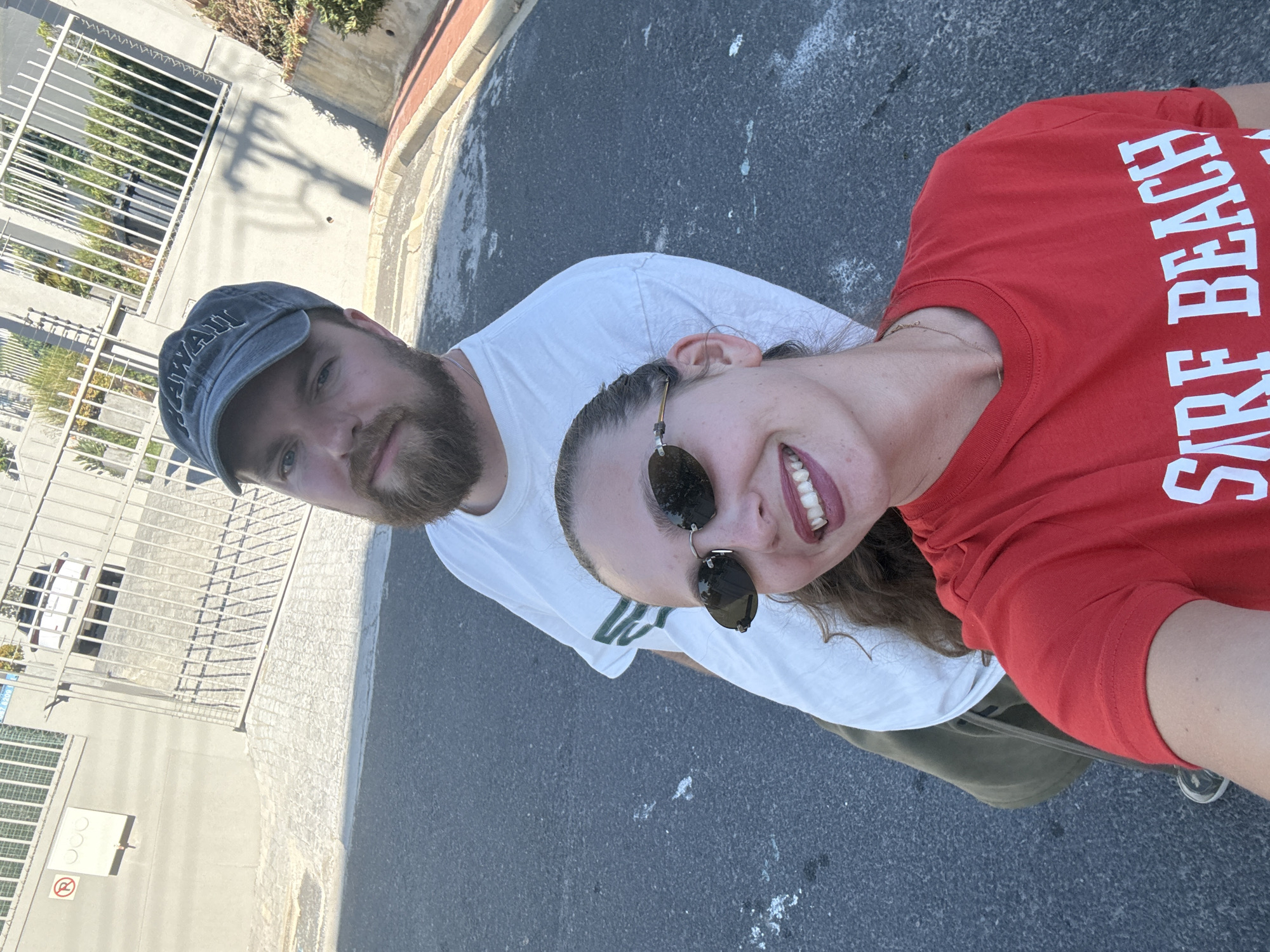 A selfie-style photograph depicts a man and a woman on an urban street in Cape Town, South Africa. The man, on the left, wears a blue baseball cap and has a beard. He is wearing a white t-shirt. The woman on the right wears sunglasses and a red t-shirt that reads "Surf Beach." They are standing on an asphalt road with some patches of white markings. In the background, there is a building with a metal fence and urban surroundings. The picture's angle suggests it was taken at eye level.
