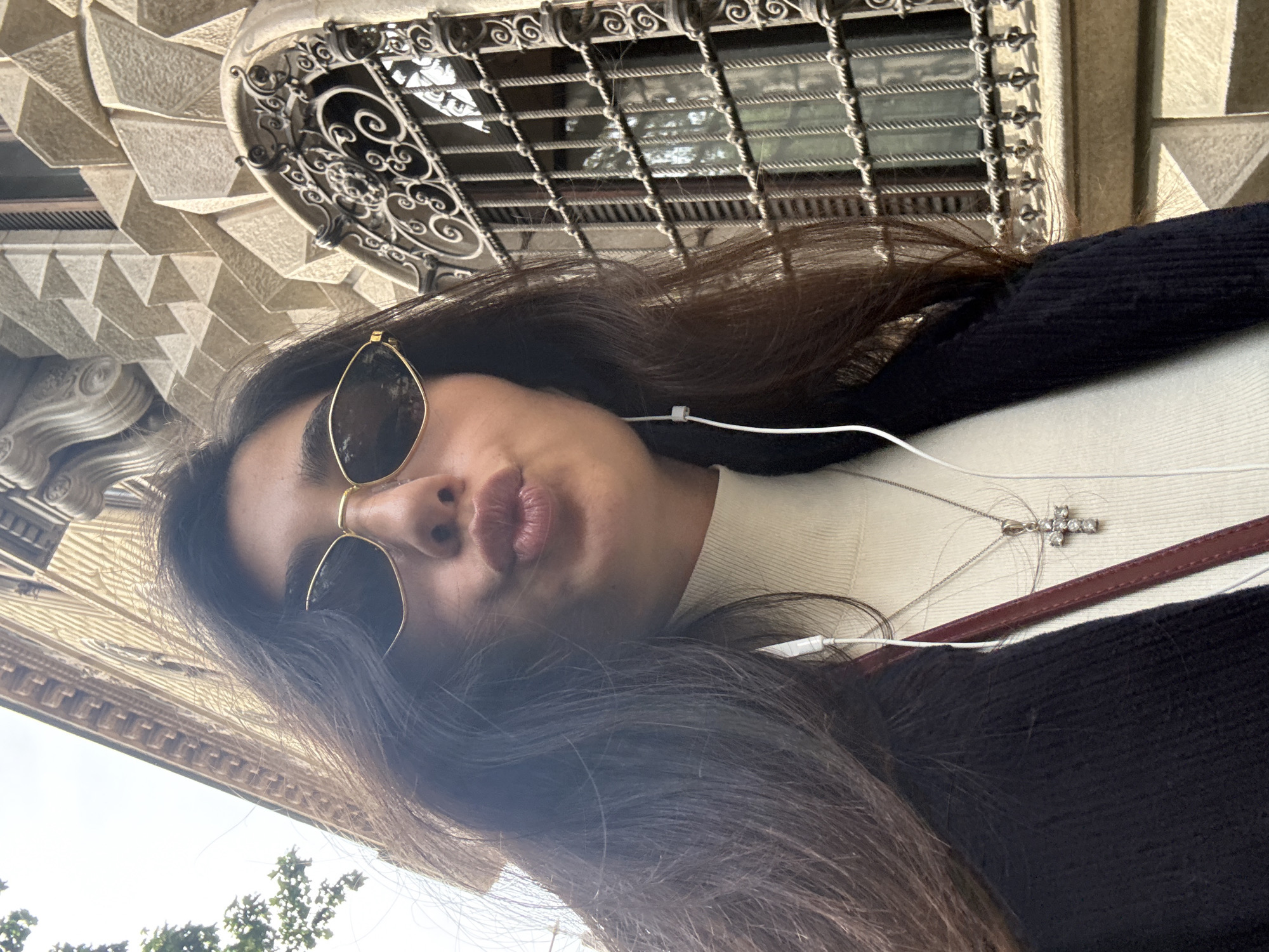 A low-angle selfie captures a young woman looking up, set against the intricate architecture of a building in Milan, Italy. The image is rotated, presenting the subject's face prominently in the center. The woman has long, dark brown hair and is wearing gold-framed sunglasses with dark lenses. Her lips are slightly puckered in a common selfie expression. She wears a light-colored, high-necked top and a dark cardigan or jacket. White wired earbuds are visible, with the wire extending from her ear. Around her neck, a silver chain holds a sparkling cross pendant adorned with small jewels.
The background showcases decorative architectural elements. Directly above the woman's head (when rotated correctly), an ornate dark metal grille or balcony railing with elaborate swirling patterns is visible. To the left, a section of the building facade features a distinctive geometric pattern of faceted, triangular stone or concrete reliefs. Further down and to the left, a more classical cornice or frieze with repeating motifs is seen, with a glimpse of an open sky and green tree tops, indicating an outdoor setting. The lighting suggests daytime, with diffused natural light illuminating the scene. No discernible text is visible in the image.