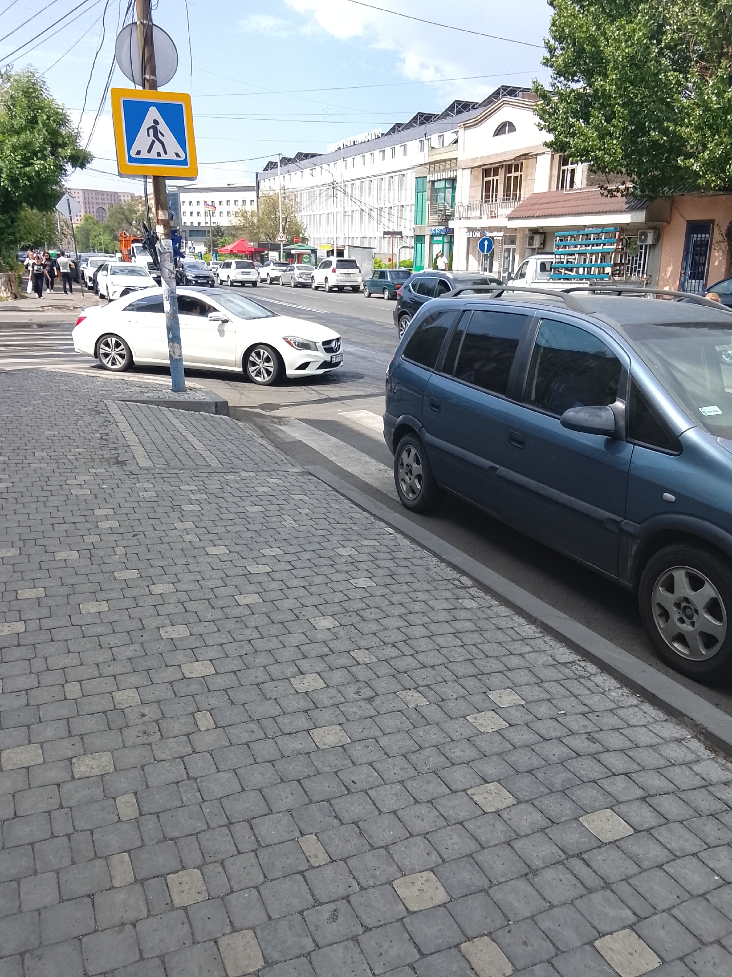 This image depicts a street scene in an urban environment during daylight hours, under what appears to be fair weather. The foreground is dominated by a grey, textured pedestrian walkway paved with interlocking rectangular stones, some of which are lighter in color. A dark grey minivan is parked along the curb on the right side of the image. Beyond the minivan, a white car is driving across a pedestrian crossing, its brake lights illuminated. The street appears to be a two-way road with multiple lanes of traffic.
In the background, a row of multi-story buildings lines the street. These buildings have a mixture of architectural styles, with some featuring balconies and arched windows, while others appear to be more modern. Trees with green foliage are visible on the left and right sides of the frame, suggesting a moderately vegetated urban area. A pedestrian crossing sign, a blue square with a yellow border and a white pedestrian icon, is prominently displayed on a utility pole. Several people are visible walking on the sidewalk on the left side of the street, suggesting a bustling area. The overall atmosphere is one of everyday urban activity. There is no discernable text in the image.