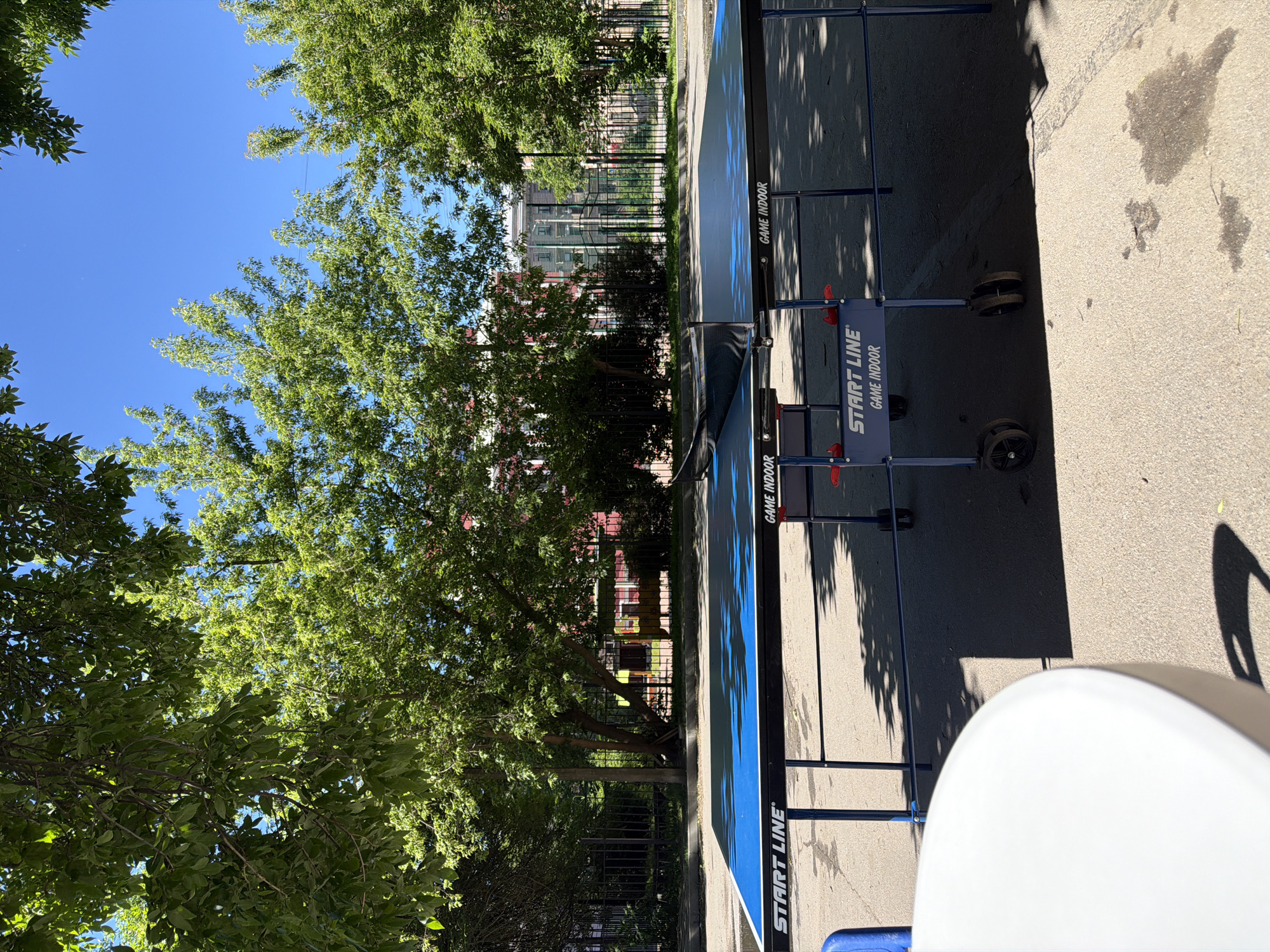 The image shows a blue and black ping pong table set up outdoors on an asphalt surface. The table has the "START LINE" logo and "GAME INDOOR" written on it. There is a net stretched across the middle of the table. The table is positioned in front of a black metal fence and a row of trees with lush green leaves. Beyond the fence, there is a building with red accents and a clear blue sky above. The overall scene suggests a recreational area within a city, likely in Saint Petersburg, Russia, given the architectural style of the building and the presence of trees and a fence commonly found in urban parks or courtyards. There are no people visible in the image. A white, round table is partially visible in the foreground, casting a shadow on the asphalt.