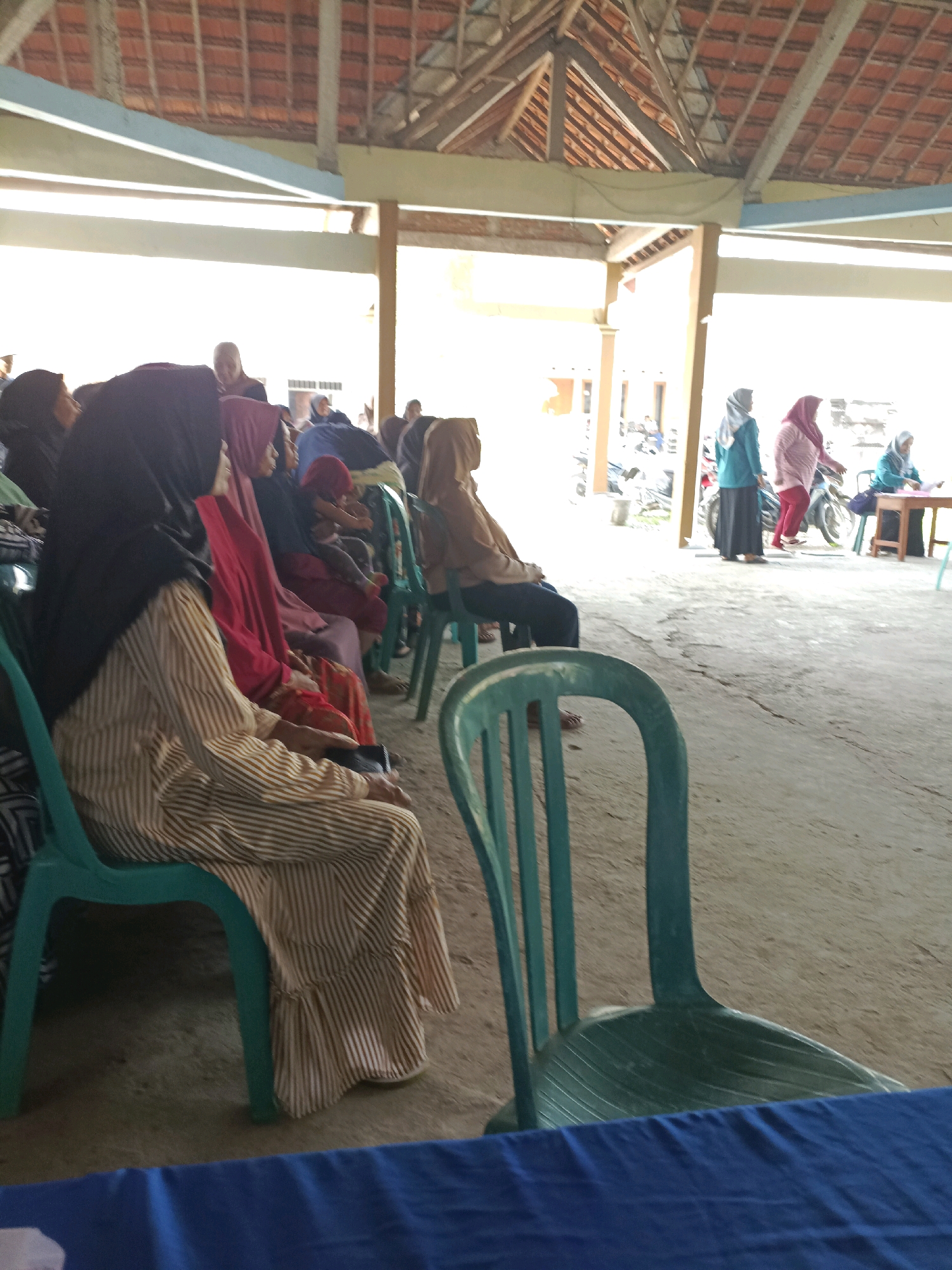 The image depicts an outdoor gathering under a covered structure with a tiled roof. In the foreground, several individuals, predominantly women wearing hijabs, are seated on green plastic chairs. A woman in a striped dress and black hijab is in the left foreground, facing away from the camera. To her right, other women in various colored hijabs and clothing are also seated. In the middle ground, more people are visible, including a child being held. Further back, several individuals stand near motorcycles, and a woman sits at a table. The ground appears to be concrete or dirt. The setting suggests a community event or meeting in Rancang Kencono, Indonesia.