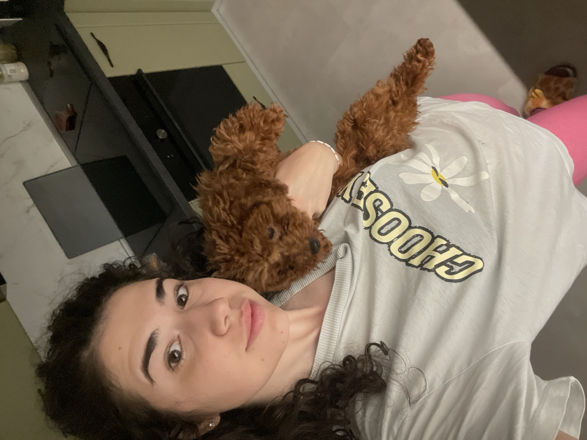 A medium shot angled slightly downward depicts a woman lying down, with a small, curly-haired brown dog lying on her chest. The woman has dark, curly hair and is wearing a light-colored t-shirt with the word "CHOOSE" printed on it in yellow letters. Her arm is positioned around the dog. The background includes a kitchen countertop and cabinets on the left side of the frame, and a pink object, possibly a cushion or exercise ball, is visible behind the woman. The location in Sofia, Bulgaria is not discernible from the image.