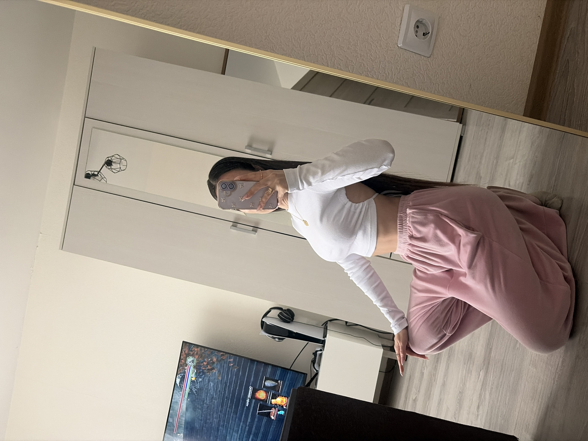 A person is reflected in a mirror, kneeling in a room. The person is holding a smartphone with a floral-patterned case. The person is wearing a white long-sleeve top and pink sweatpants. In the background, there is a wardrobe with a mirror, a television displaying a video game, headphones, and a game console. The room is located somewhere in Saint Petersburg, Russia. An electrical outlet is visible on the wall near the top of the mirror frame.