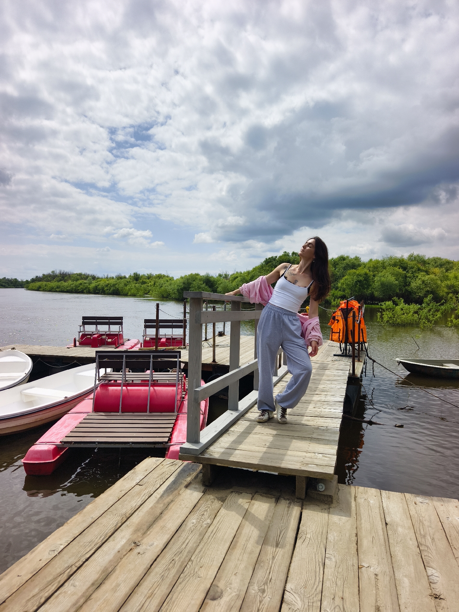 A full shot captures a woman standing on a wooden pier near the city of Onokhino, Russia. She is wearing a white tank top, grey sweatpants, and a pink open-front jacket. Her head is tilted back, and her eyes are closed, with her left arm resting on a railing. In the background, the wide, calm river is lined with lush green trees. Several red paddle boats are docked alongside the pier, as well as two white boats. Two orange life vests are stacked on a post further down the pier. The sky above is filled with dramatic, grey and white clouds, suggesting an overcast day. The wooden planks of the pier are visible in the foreground, showing signs of wear and weathering.