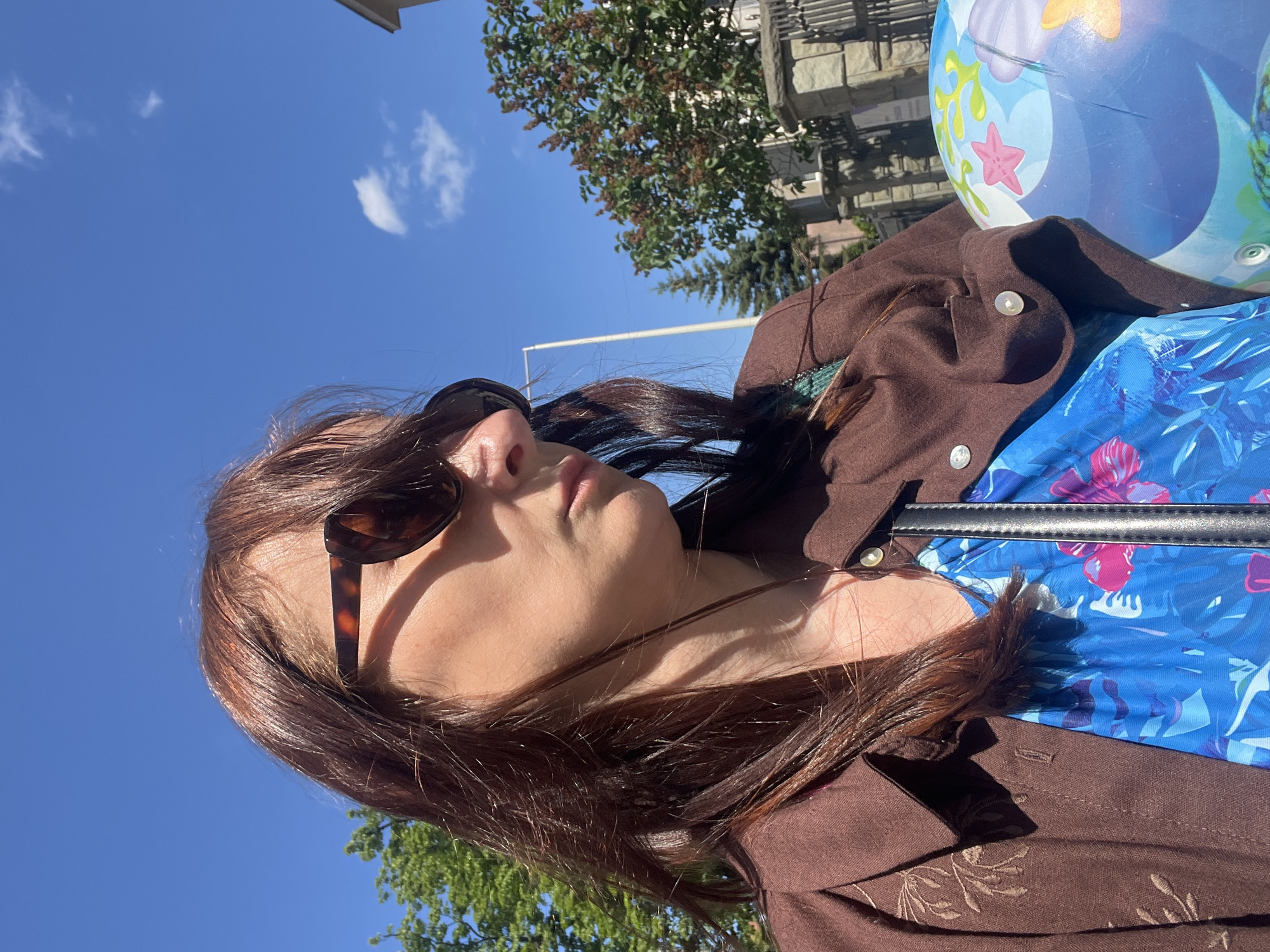 A female subject is captured in a medium close-up, low-angle, outdoor photograph taken on a clear, sunny day. She possesses dark brown, shoulder-length hair and wears dark brown tortoiseshell sunglasses. Her attire includes a brown collared shirt or jacket featuring light-colored buttons and embroidered leaf patterns on the left lapel, layered over a vibrant blue garment with a tropical floral pattern in shades of blue and pink. A dark strap, likely from a bag, is visible across her chest. The subject holds a blue, partially inflated beach ball adorned with starfish and underwater imagery. The background consists of a bright blue sky with sparse white clouds. Visible environmental elements include green deciduous trees on the left, a stone wall with dark metal fencing on the right, and behind it, a tree with reddish-brown foliage and a portion of a light-colored building's roof. A light-colored utility pole stands in the middle ground. The scene is illuminated by strong sunlight, creating distinct shadows. The location is an unspecified outdoor setting, possibly urban or suburban.