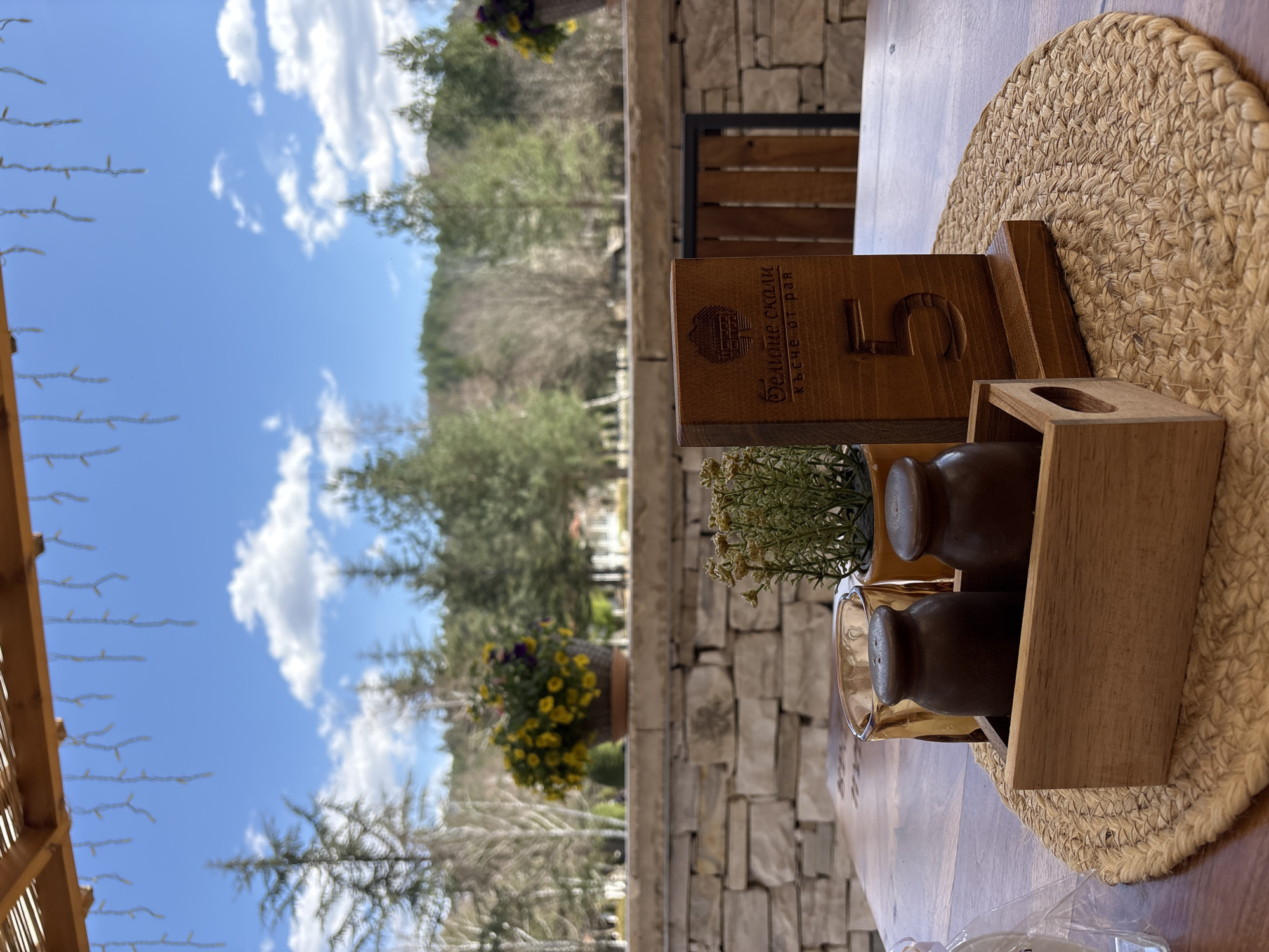 Outside, in Koprivlen, Bulgaria, a wooden table with a round placemat and condiment holders is set. A wooden table number is visible, and greenery is arranged. In the background, trees, a building, and a blue sky with clouds are visible.
