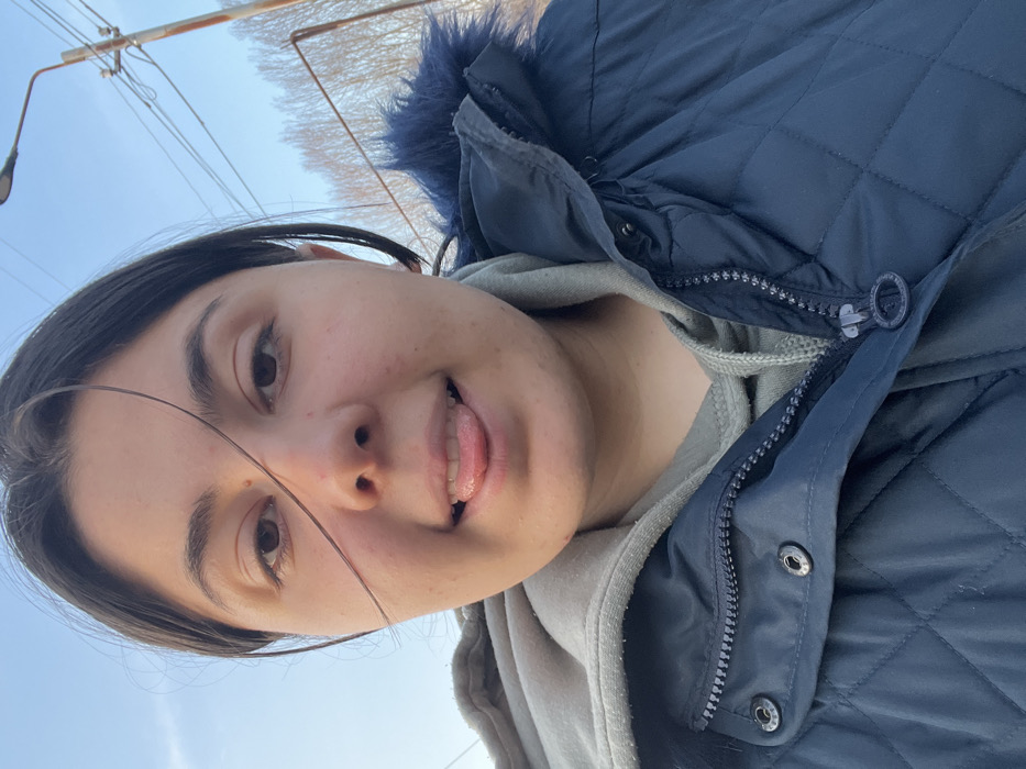 The image displays a close-up, chest-up outdoor portrait of a young adult female in Talin, Armenia. She has dark hair, a medium complexion, and dark eyes. A single strand of dark hair crosses her face over her right eye and nose, and a small, dark mole is visible above her right eyebrow. Her mouth is open, and her tongue is extended forward. She is dressed in a light gray hooded sweatshirt, layered under a dark blue quilted winter jacket. The jacket features a visible metallic zipper, partially unzipped, and metallic snap fasteners on the front placket. A dark blue faux fur trim is discernible around the jacket's hood/collar area on the subject's right side. The background consists of a clear, light blue sky, indicating daylight. To the upper right, a utility pole supports multiple utility lines and an attached street light fixture. In the distant background, a line of deciduous trees or shrubs, devoid of leaves, suggests a colder season.