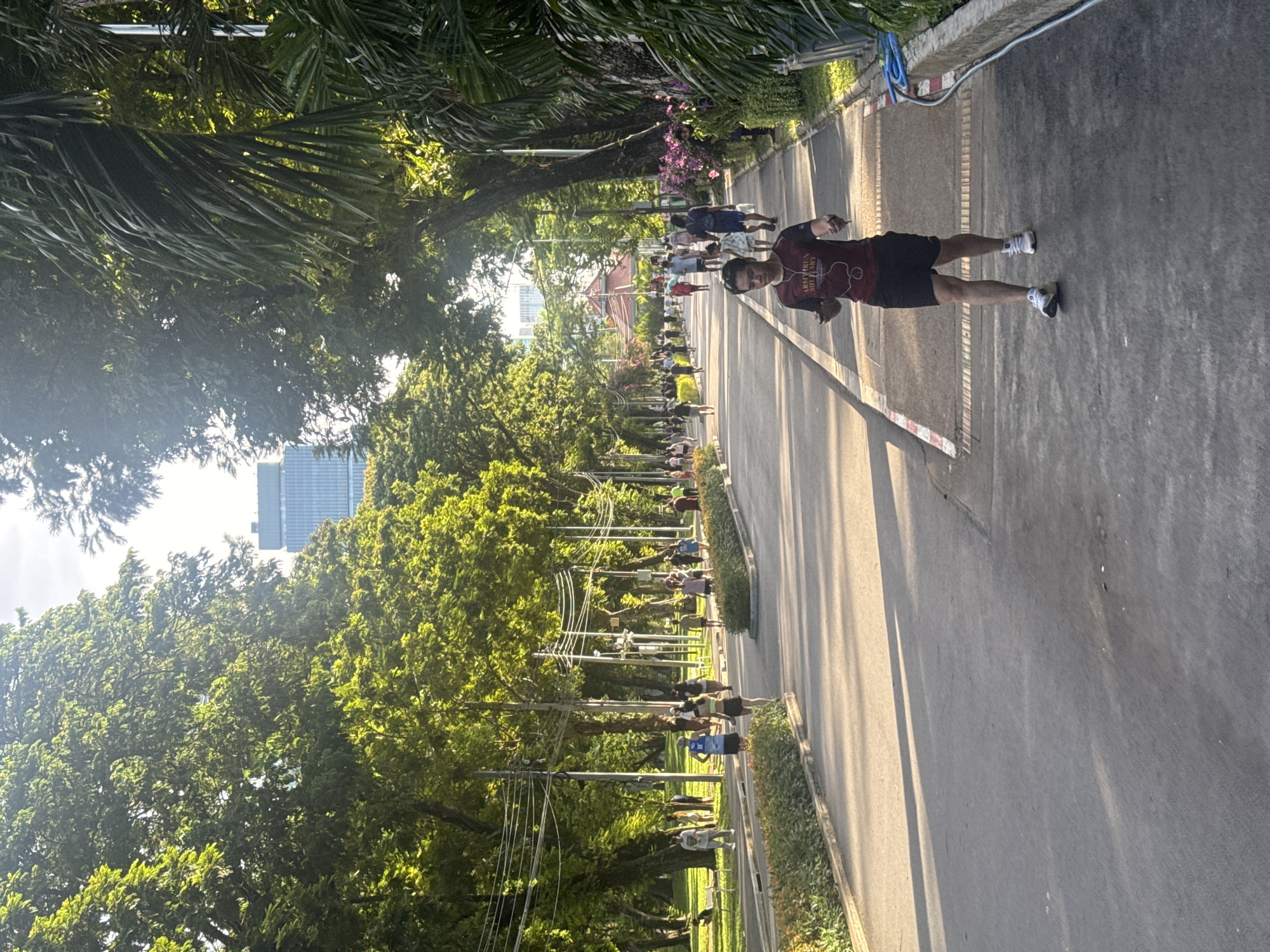 A long, tree-lined road stretches into the distance in this outdoor scene. A man in a dark shirt and shorts is jogging towards the camera on the right side of the road. A group of people are walking or jogging further down the road. Trees line both sides of the road, and a tall building can be seen in the distance. Overhead power lines are also visible. The location is likely a park or recreational area in Bangkok, Thailand, based on the urban setting and tropical vegetation.