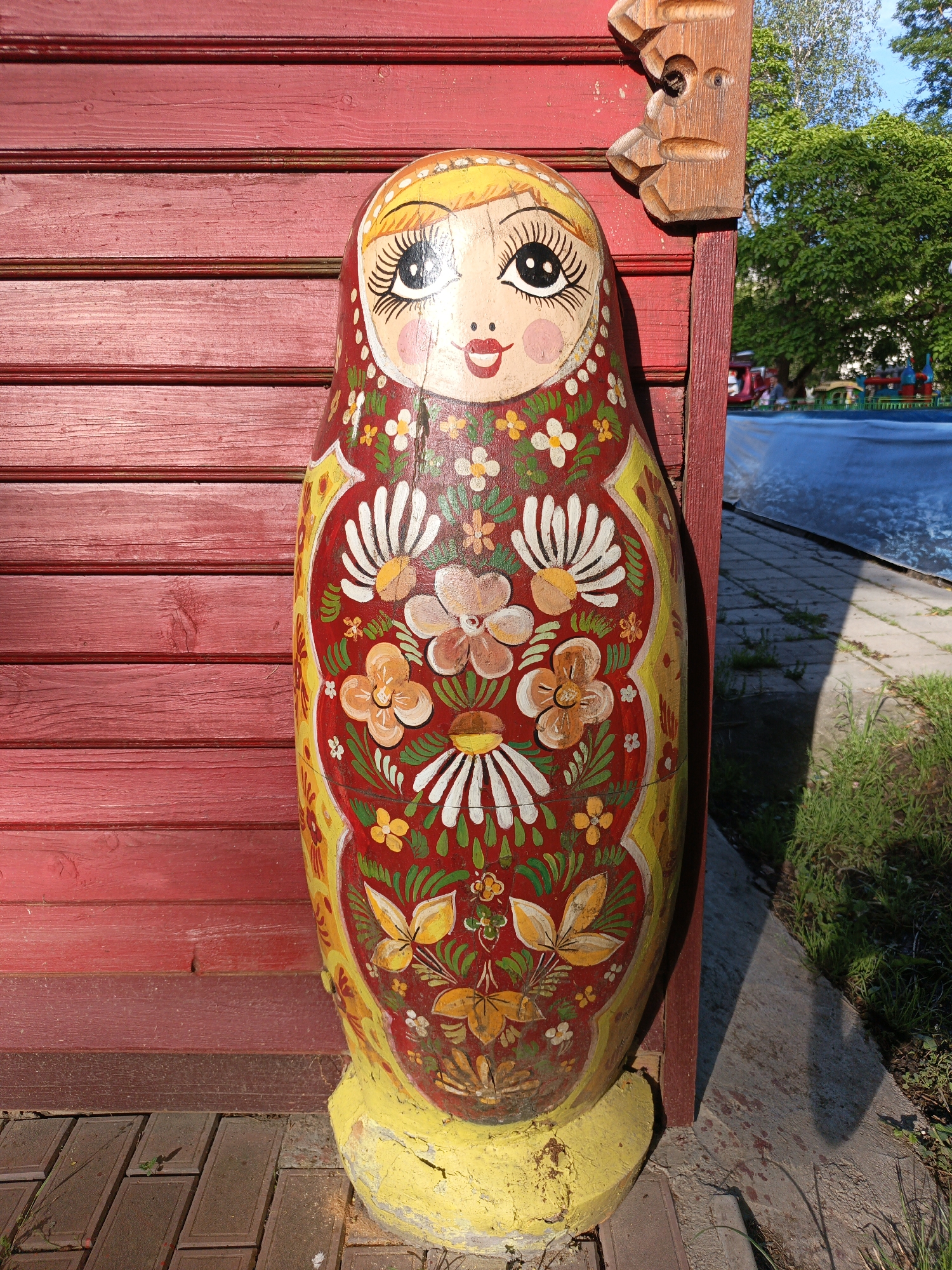 A large, colorful matryoshka (Russian nesting doll) statue stands prominently in an outdoor setting. The doll is painted in vibrant red and yellow, adorned with intricate floral patterns in white, orange, and green. It features a cheerful face with yellow hair, large wide-open eyes with long black lashes, rosy cheeks, and red smiling lips. The doll shows some signs of wear, including visible cracks on its face and chipped paint on its yellow, concrete base.
Behind the statue is a weathered red wooden wall composed of horizontal planks. To the right of the doll's head, a carved wooden decoration extends from the red wall. Beyond the wall and to the right, a bright blue sky is visible above lush green trees. Below the trees, a large blue tarp or cover stretches over an unseen structure, possibly a pool or an enclosed attraction, with distant blurred shapes suggesting an amusement park area and a faint human figure.
The ground around the doll consists of brown rectangular paving stones on the left and a mix of concrete, dirt, and green grass on the right, where strong shadows are cast. The scene is brightly lit by natural sunlight, indicating it is daytime. The overall ambiance is cheerful and festive, placing the statue in what appears to be a public park or recreational area in Goryachiy Klyuch, Russia. There is no visible text in the image.