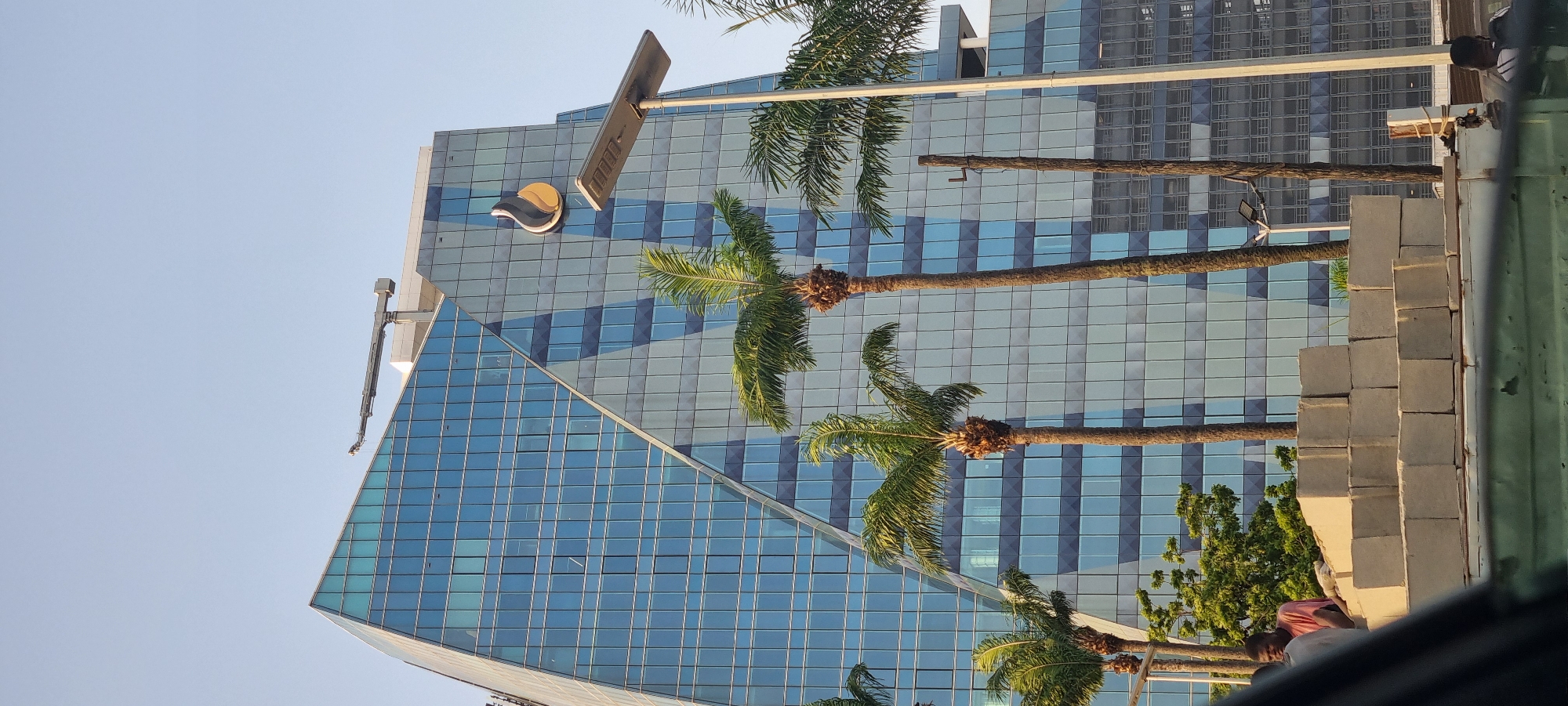 The image captures a view of a modern building in Ikoyi, Nigeria, characterized by its glass facade with a grid-like pattern. A distinctive logo is visible on the building. Several palm trees are positioned in front of the building, obscuring parts of the facade. A stack of light-colored, possibly concrete, blocks is positioned on the right side of the frame, partially obscuring the view and suggesting nearby construction. The sky is light blue, indicating clear weather. Part of a vehicle's interior is visible in the lower right corner of the image.