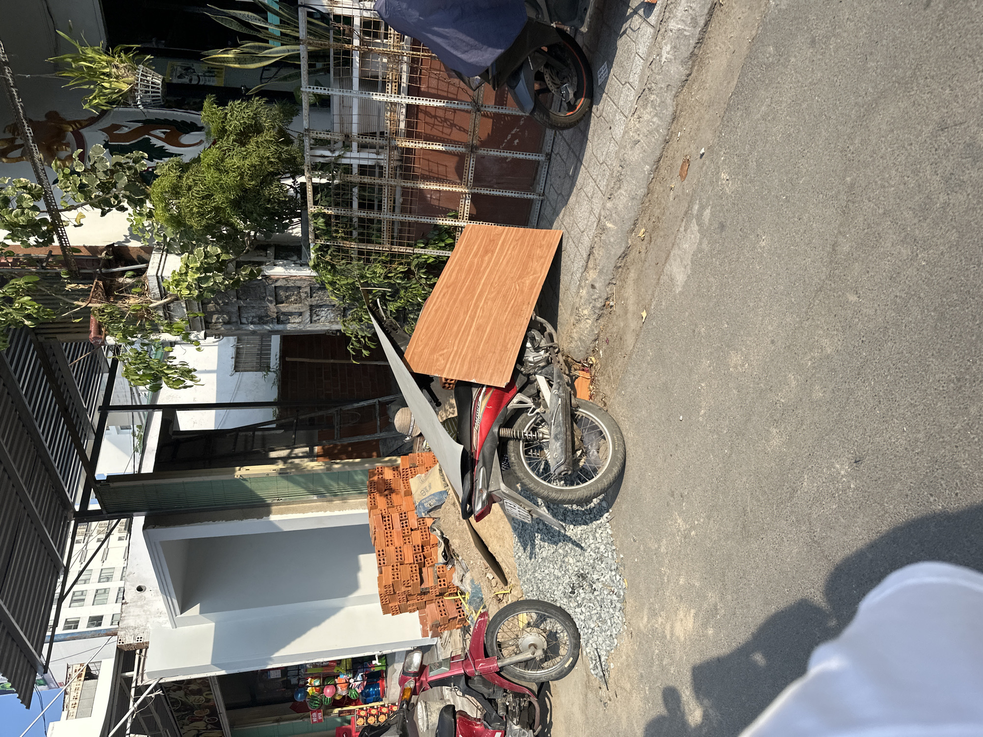 The image depicts a street scene in Bac Nha Trang Ward, Vietnam. A red and silver motorcycle is parked on the side of the road, carrying a large, rectangular piece of wood. The motorcycle is adjacent to a pile of red bricks and a small area covered in gravel. Another red motorcycle is parked nearby. In the background, there are buildings, including a shop with various items on display. Vegetation, including trees and potted plants, is visible along the building facades. The street surface is asphalt. A person, partially obscured, is visible in the lower right corner of the frame. A motorcycle is parked beside a metal gate in the background.