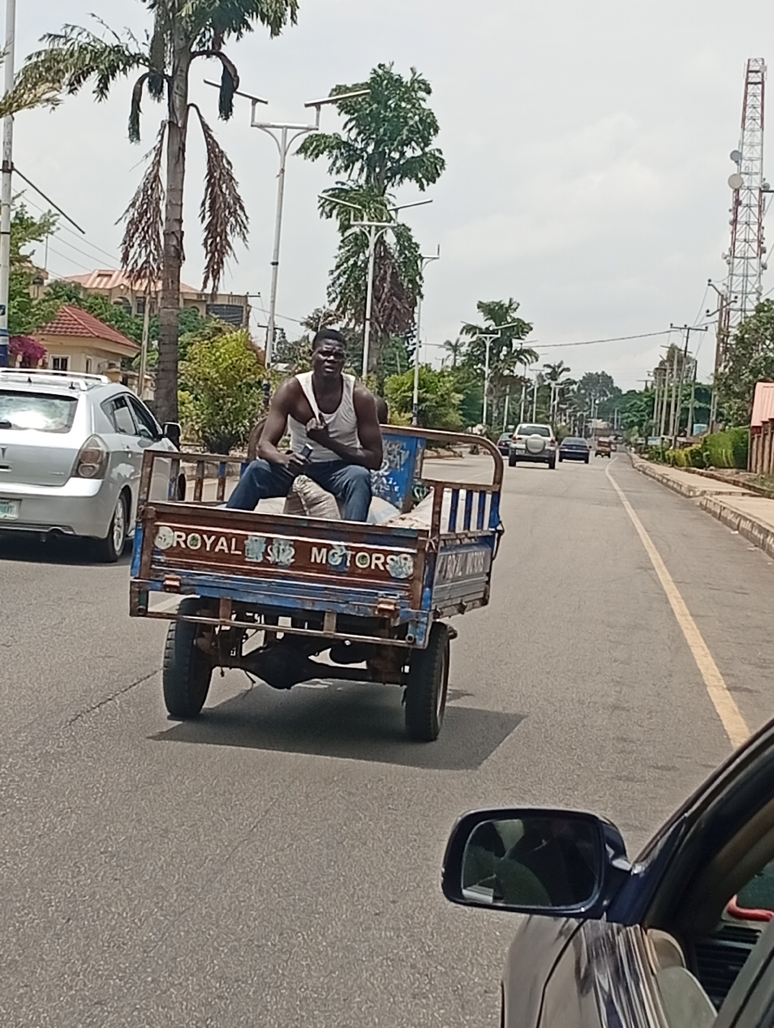 The image shows an unpaved utility vehicle on a paved roadway in Jos, Nigeria. The vehicle has signage that reads "ROYAL MOTORS". The vehicle is being occupied by a dark-skinned, dark-haired male wearing a white tank top and dark blue jeans. The male is sitting, facing forward, and appears to be holding something in his right hand. The roadway has a yellow centerline. Other vehicles and palm trees are also present in the frame, with a telecommunications tower in the distance to the right. The view is from the perspective of another vehicle, as the wing mirror of a blue sedan is visible in the lower right.