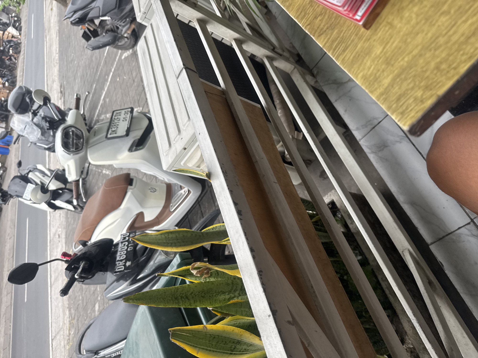 In Kuta Selatan, Indonesia, a street scene unfolds with several motorcycles and scooters parked on a tiled sidewalk. In the foreground, a railing and a potted plant with spiky, variegated leaves partially obscure the view. Beyond the railing, a white scooter with a brown seat is prominent, its license plate reading "DK 3892 FDX". Adjacent to it, a black scooter with a similar license plate, "DR 6805 CD", is also visible. Further down the street, more motorcycles and scooters are parked, some covered with tarps. The background shows a road with some traffic. A wooden table with a stack of red-and-white items sits in the lower right corner of the frame, and a person's leg is visible in the bottom center.