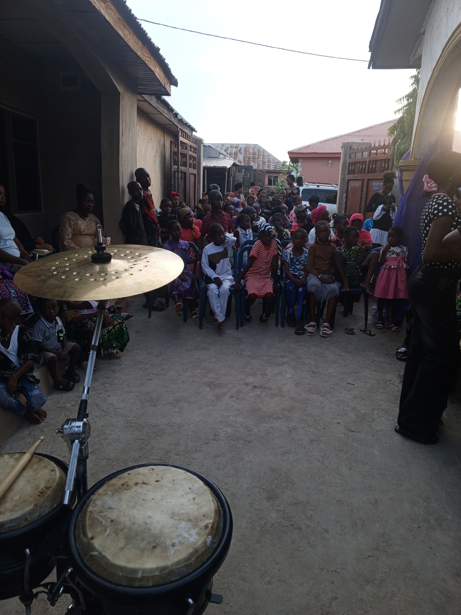 A gathering of people, mostly children, are seated on chairs in an outdoor setting with buildings visible in the background. A set of bongo drums and a cymbal stand in the foreground. The location is in a city and country marked as Unknown, Unknown.