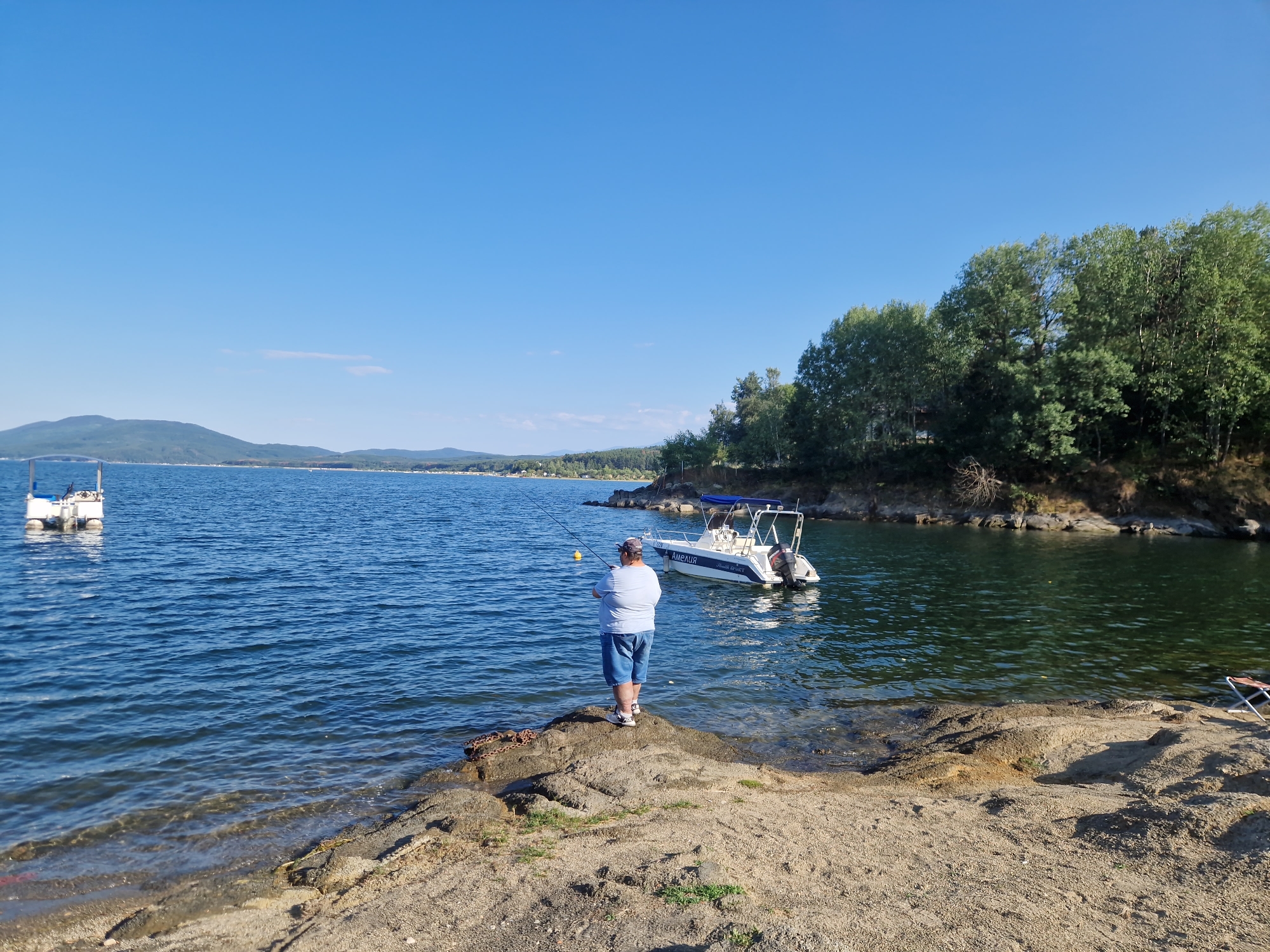 The image captures a serene lakeside scene in Dolni Pasarel, Bulgaria. The foreground features a sandy shoreline with rocky outcrops, leading to the calm, blue waters of a lake. In the middle ground, a person stands on a rock, fishing with a rod. The person is wearing a light blue t-shirt and blue shorts. A white motorboat with a blue canopy is anchored nearby in the water, with the name "АМЕЛИЯ" visible on its side. The background is dominated by a lush, green forest that slopes down to the water's edge, with rolling hills and mountains visible in the distance under a clear blue sky. There is also a white floating structure visible on the left side of the lake, possibly a marker or a small dock. The overall atmosphere is peaceful and idyllic.