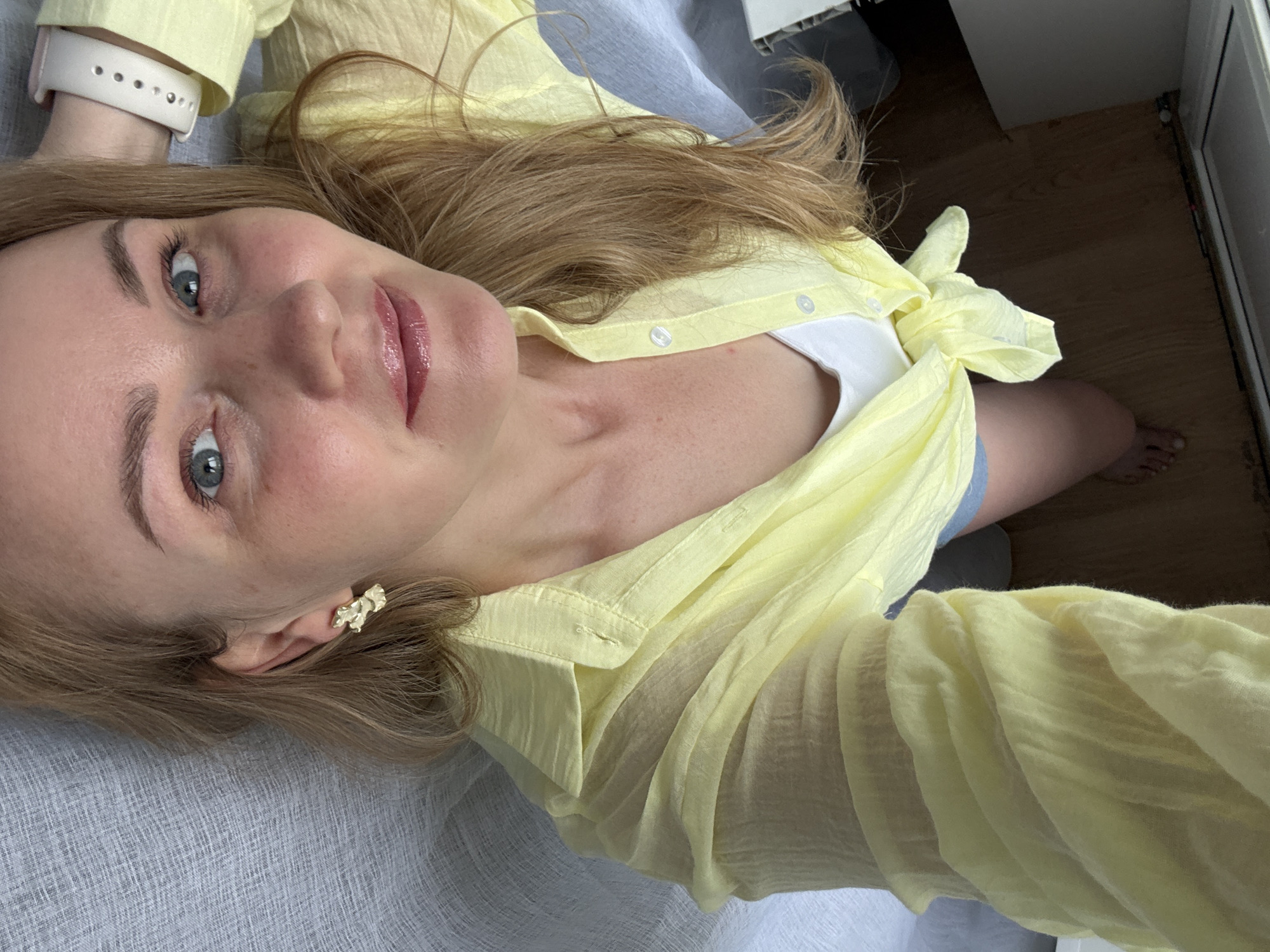 A woman with blonde hair and blue eyes is lying on a textured grey surface. She is wearing a light yellow, loosely buttoned shirt tied at the waist, revealing a white tank top. A small gold earring is visible in her left ear. Her right hand is resting behind her head, and she is wearing a white watch on her left wrist. The background consists of a grey textured fabric and a wooden floor. The scene appears to be indoors. There are no discernible landmarks or features indicating the specific location in Chaikovsky, Russia.