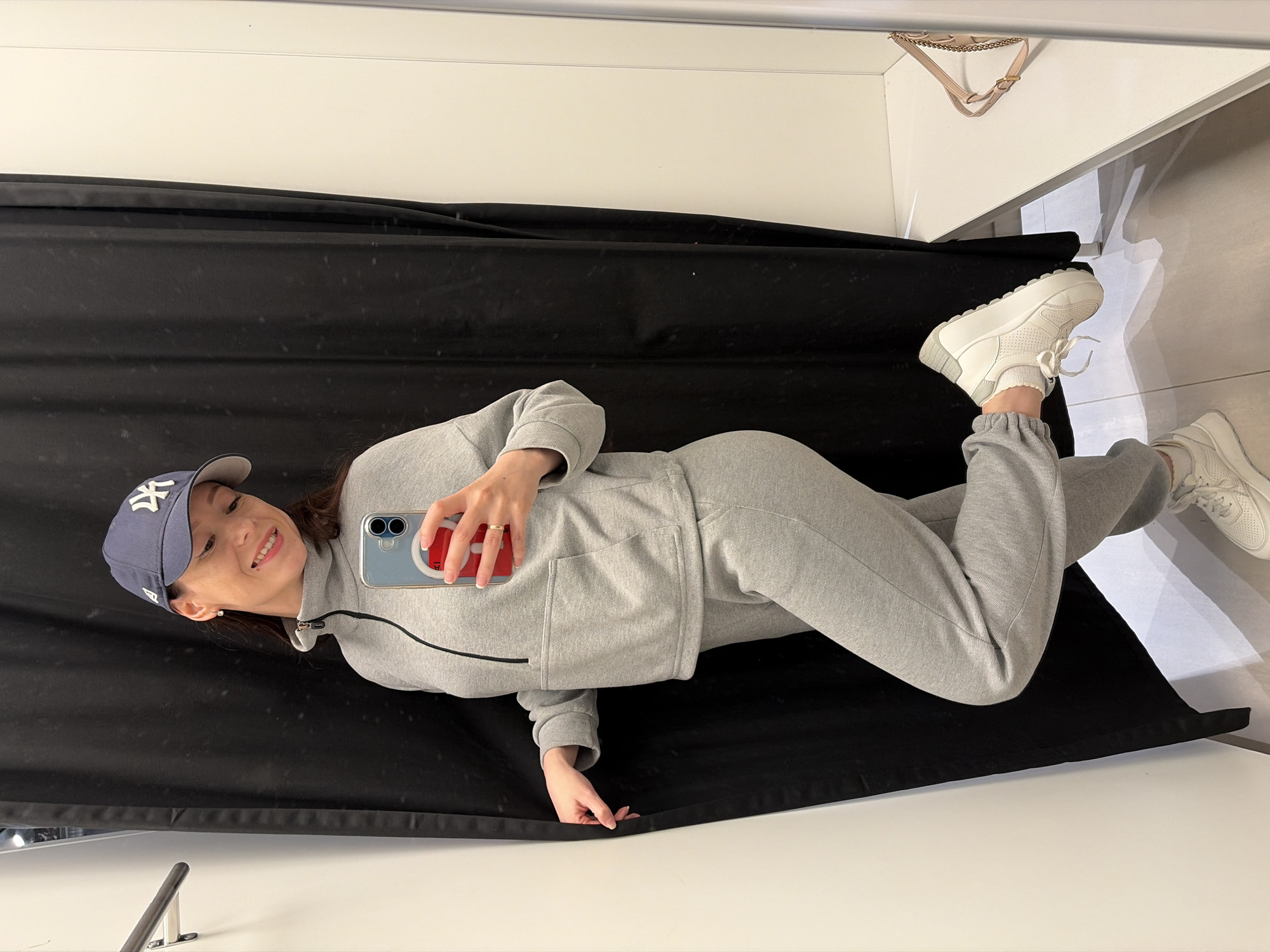 A young woman takes a mirror selfie, captured in a rotated, almost horizontal orientation. She is dressed in a casual grey tracksuit, comprising a zippered hoodie and matching sweatpants, paired with chunky white platform sneakers and white ruffled socks. A dark blue baseball cap with a prominent white "NY" logo covers her head, and she smiles broadly, looking towards the camera in her hand. She holds a smartphone featuring a light blue case with a red attachment on the back.
The setting appears to be an indoor space, likely a changing room or a retail store, indicated by the dark black curtain serving as a backdrop behind her. The foreground and side show clean, bright white surfaces, possibly a floor and a counter or bench, creating a reflective environment consistent with a mirror shot. In the upper part of the reflection, a light-colored handbag with a chain strap is visible. The image conveys a cheerful and relaxed mood, highlighting her outfit. The location context for the image is Япрыково, Russia.