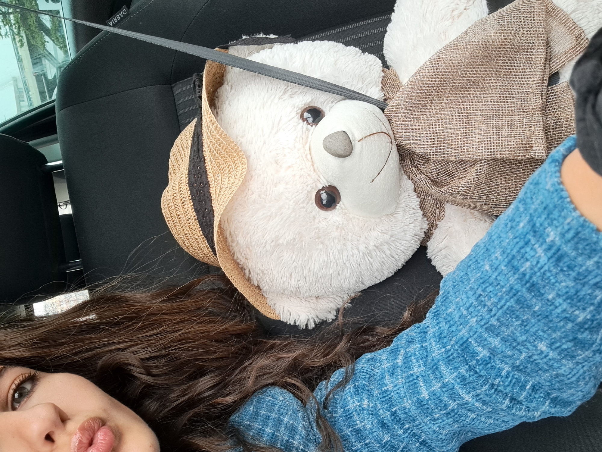 A person is in the passenger seat of a car with a teddy bear beside them. The teddy bear is white and wearing a straw hat. A seatbelt is across the teddy bear. The person is wearing a blue sweater. The car seat is black. The photo appears to be taken in Sofia, Bulgaria, although no specific landmarks are clearly visible.