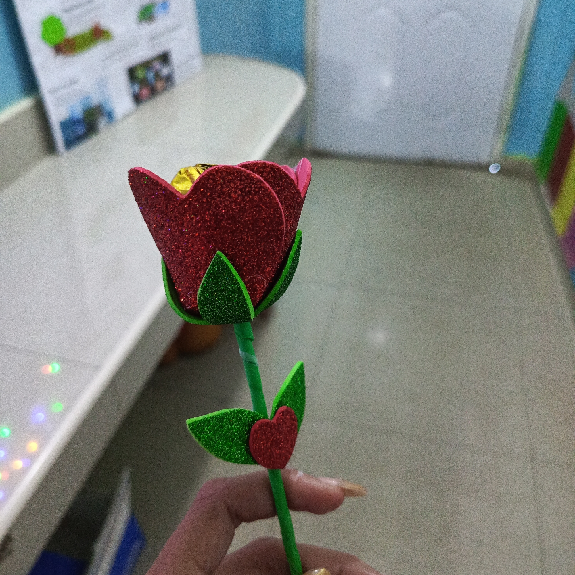 A hand holds a red glittery foam rose, with green glittery foam leaves and a red glittery foam heart. The rose appears to have a gold-wrapped candy inside. The location is indistinct, with parts of a table, a bulletin board, and a tiled floor visible. There are no clear identifying features to definitively place the image in Juan Griego, Venezuela.