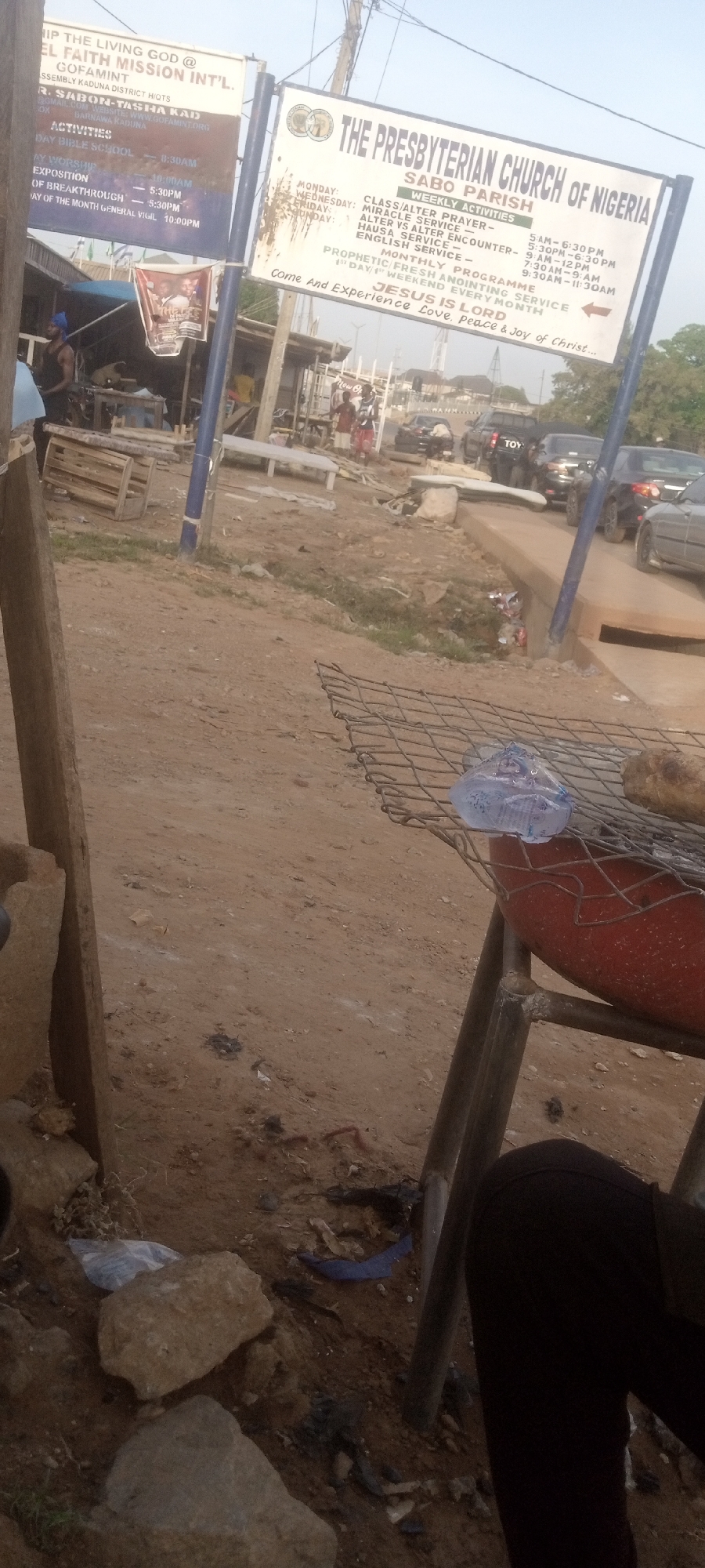 The image shows a street scene in Yelwa, Nigeria. In the background, there are two church signs. One sign reads "THE LIVING GOD @ EL FAITH MISSION INT'L" and the other reads "THE PRESBYTERIAN CHURCH OF NIGERIA." There are people walking on the street, and cars are driving. The foreground is focused on the area surrounding what appears to be a grill made from an old red propane tank. There is trash on the ground, including plastic bottles and bags, and some rocks are strewn about. A person's leg can be seen in the lower right corner.